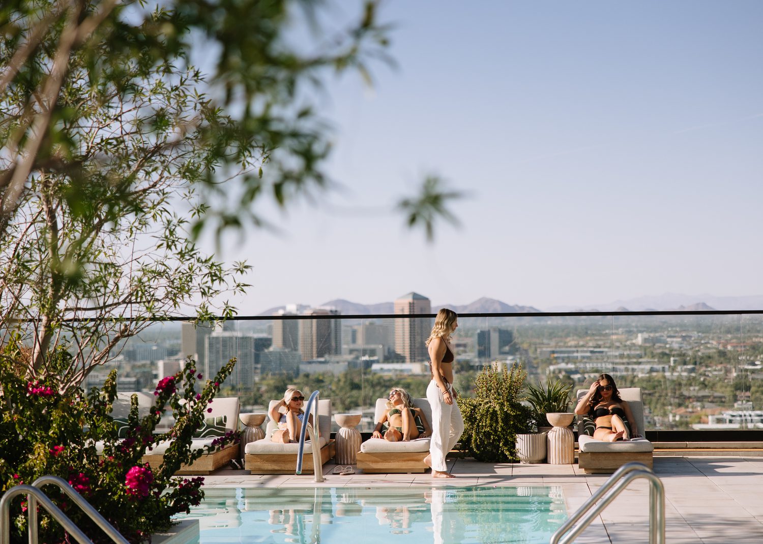 Four women relax by a rooftop pool at New Student Apartments ASU, with city buildings and mountains in the background. Two lounge on chairs, one stands in swimwear, and flowers frame the scene under a clear blue sky.
