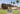 Four young women walk together on a sunny college campus lined with palm trees, strolling past modern and historic buildings and discussing their favorite Rambler 3x3 floorplans.