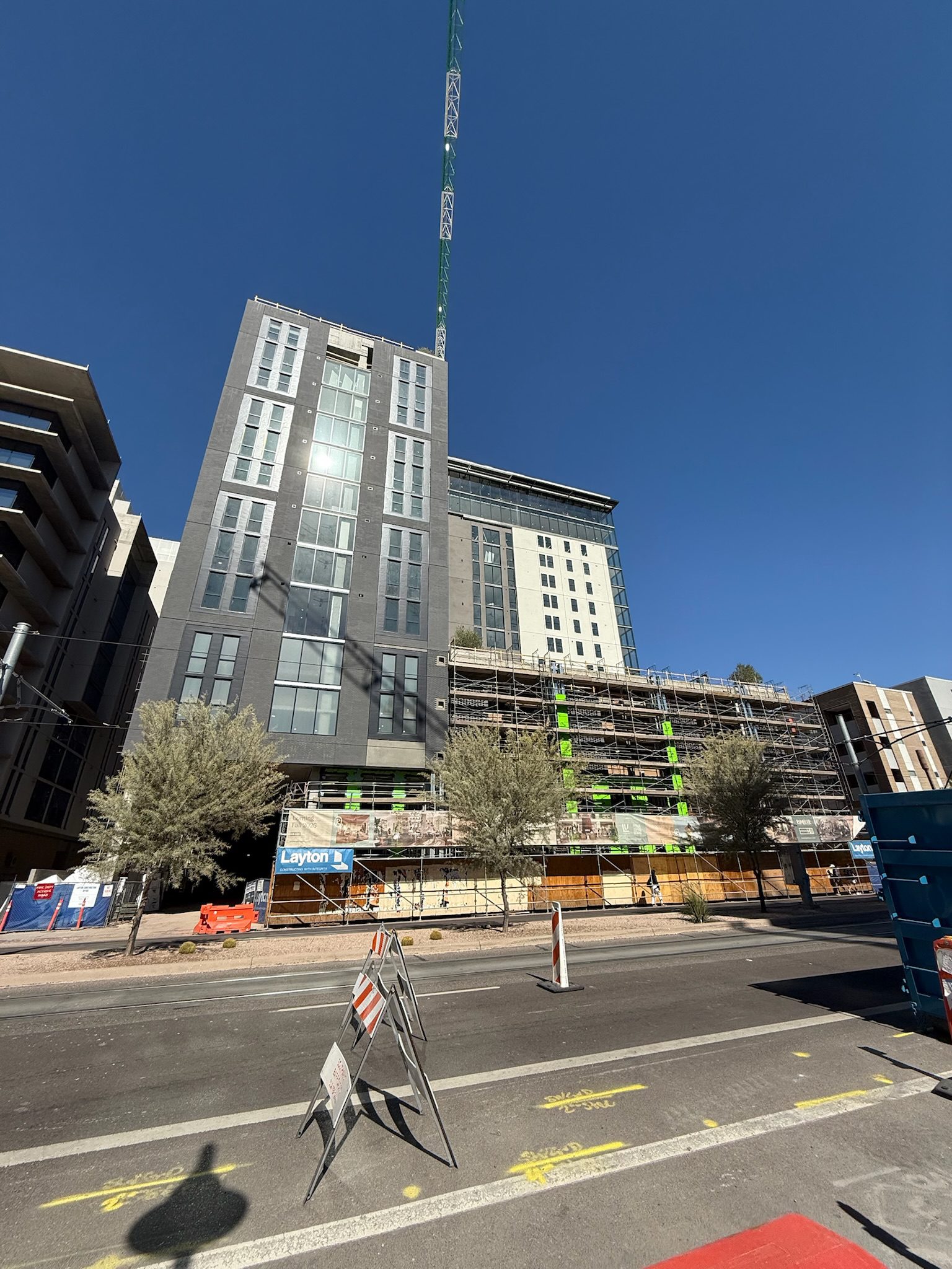 A tall building under construction with scaffolding and green insulation panels on the lower floors, a crane above, and traffic barriers and cones on a street in front under a clear blue sky offers a dynamic construction update.