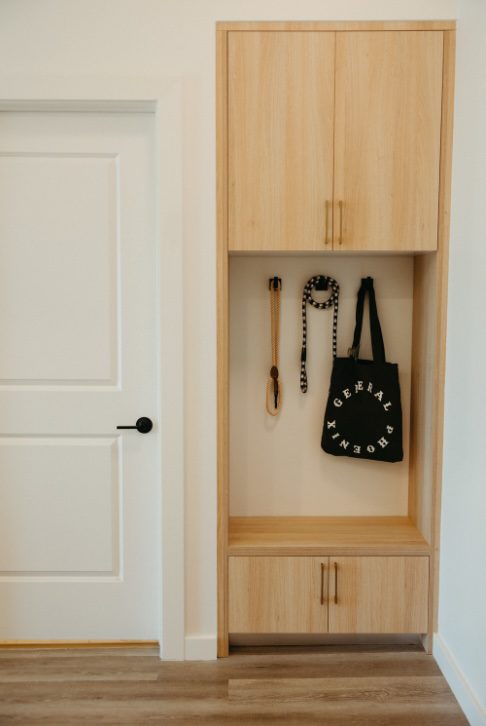 A light wood built-in entryway with cabinets, hooks holding a black tote bag with white text, a black and white lanyard, and a dog leash. A white door with a black handle is to the left.