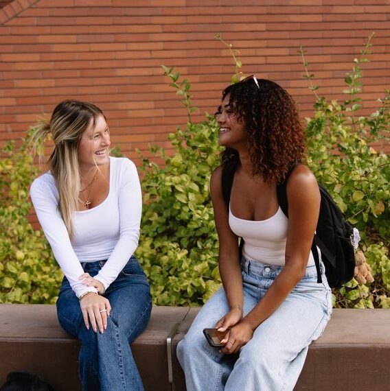 Two young women sit on a low concrete wall outside, smiling and talking. Both wear white tops and jeans, with backpacks beside them. Green bushes and a brick wall are in the background.