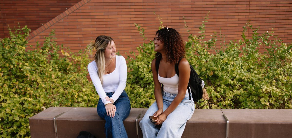 Two young women sit on a low concrete wall outside, smiling and talking. Both wear white tops and jeans, with backpacks beside them. Green bushes and a brick wall are in the background.