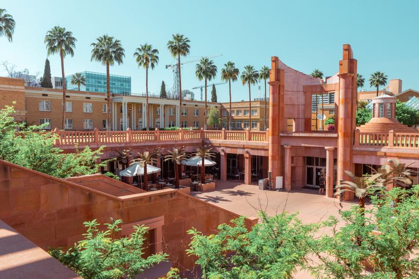 A sunlit courtyard at Arizona State University features red sandstone architecture, palm trees, outdoor seating with umbrellas, and academic buildings in the background under a clear blue sky.