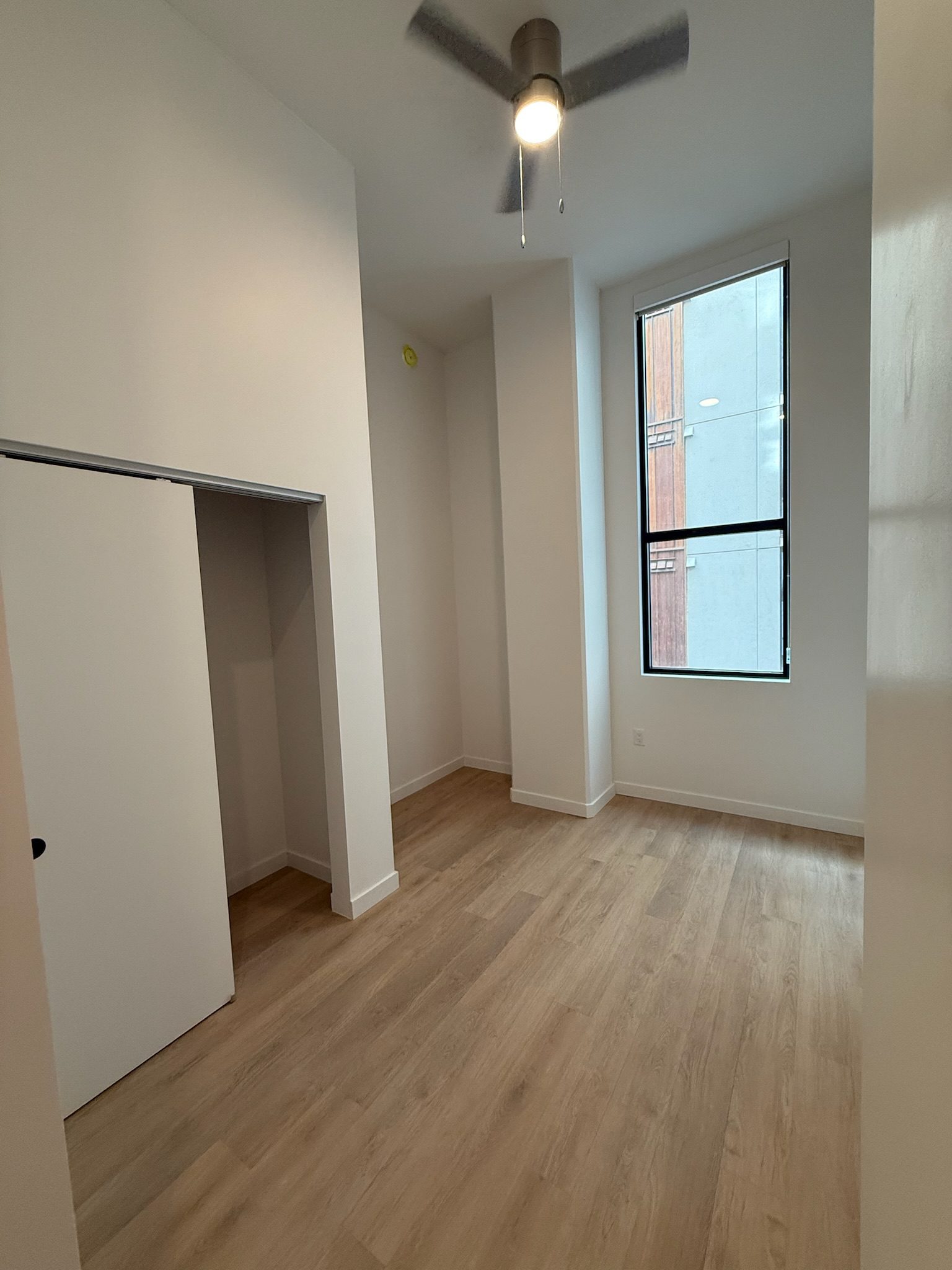 A small, empty room with light wood flooring, white walls, a ceiling fan with light, a tall window, and an open closet space with sliding doors. This construction update highlights the natural light entering from the window.