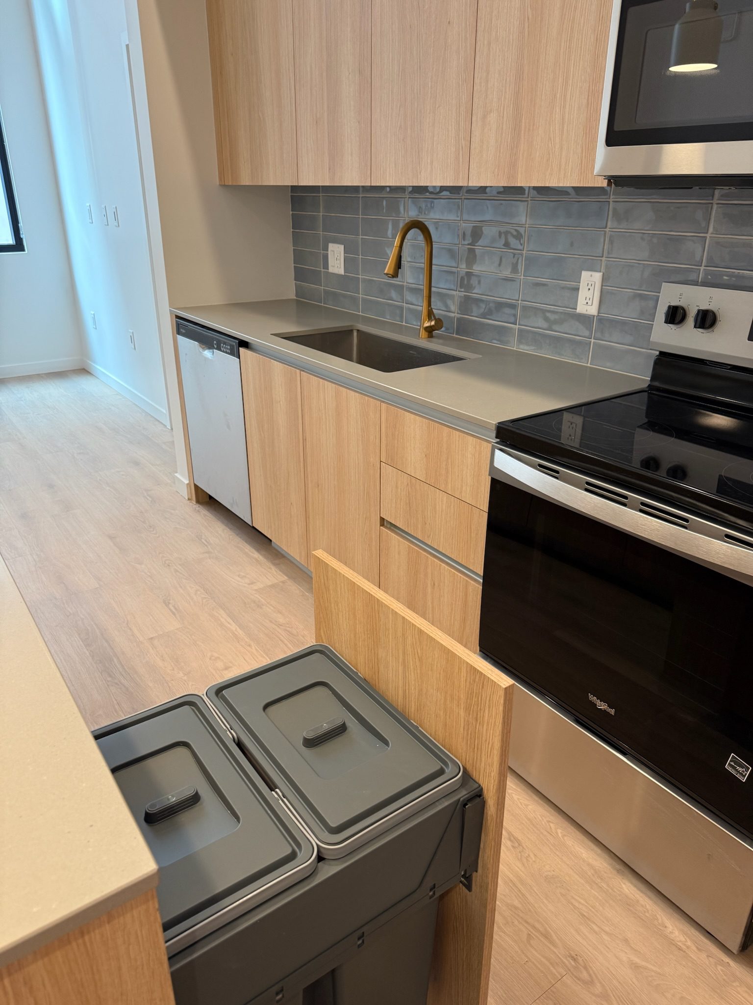 A modern kitchen with light wood cabinets, gray countertops, and a gold faucet. The cabinet under the counter is open, revealing two gray trash bins. Stainless steel appliances and a blue-tiled backsplash are visible.