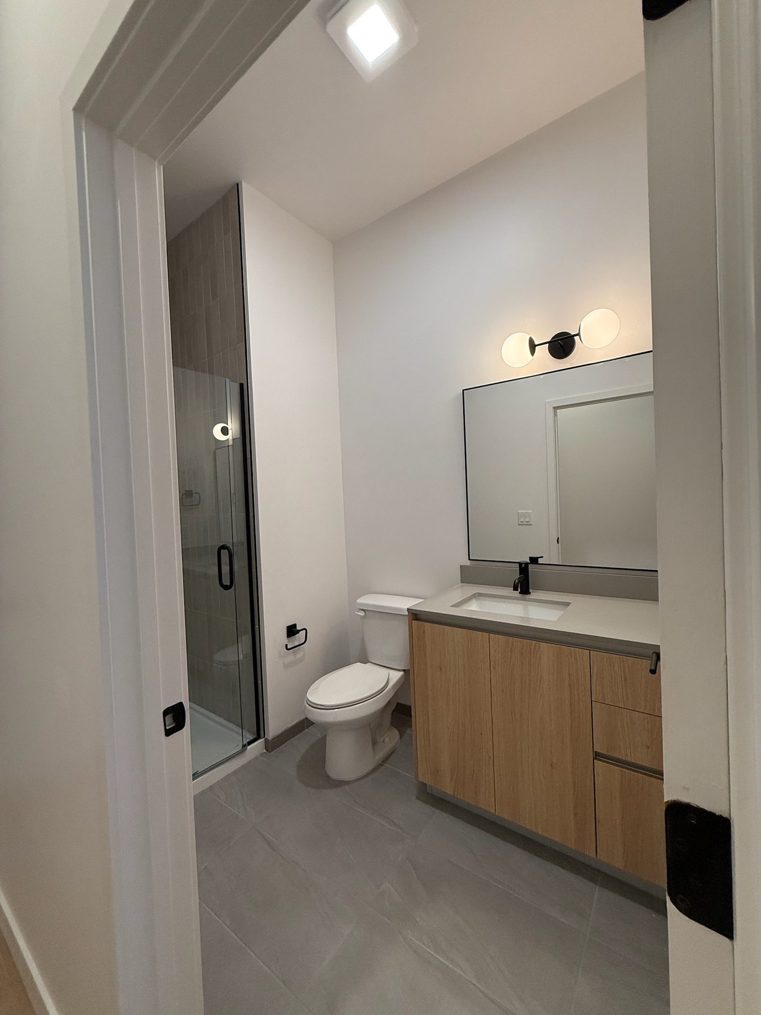 A modern bathroom with a glass-enclosed shower, toilet, wooden vanity with a sink, large mirror, and dual globe wall light. The room has light gray floor tiles and white walls.