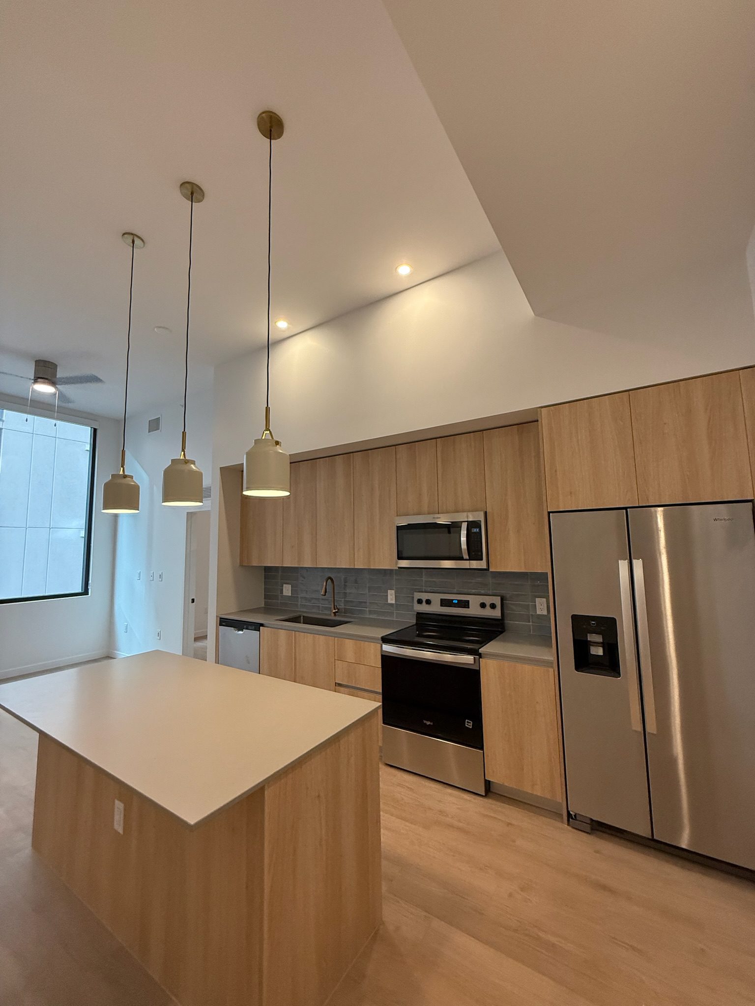 A modern kitchen with light wood cabinets, a central island, pendant lights, stainless steel appliances including a refrigerator, oven, and microwave, and a large window providing natural light.