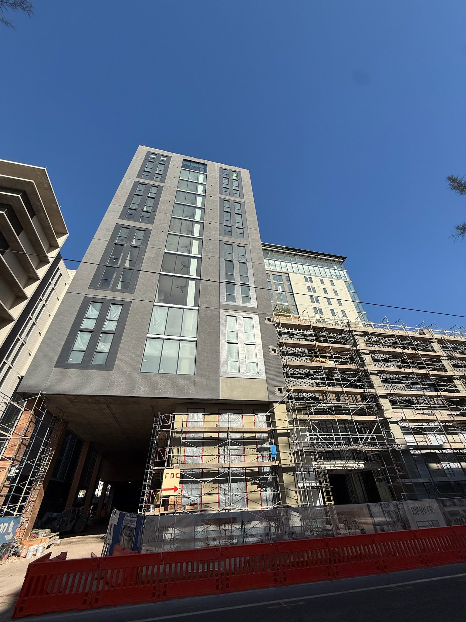 A tall modern building under construction, with scaffolding along the lower floors and a red barrier in front. The sky is clear and blue.