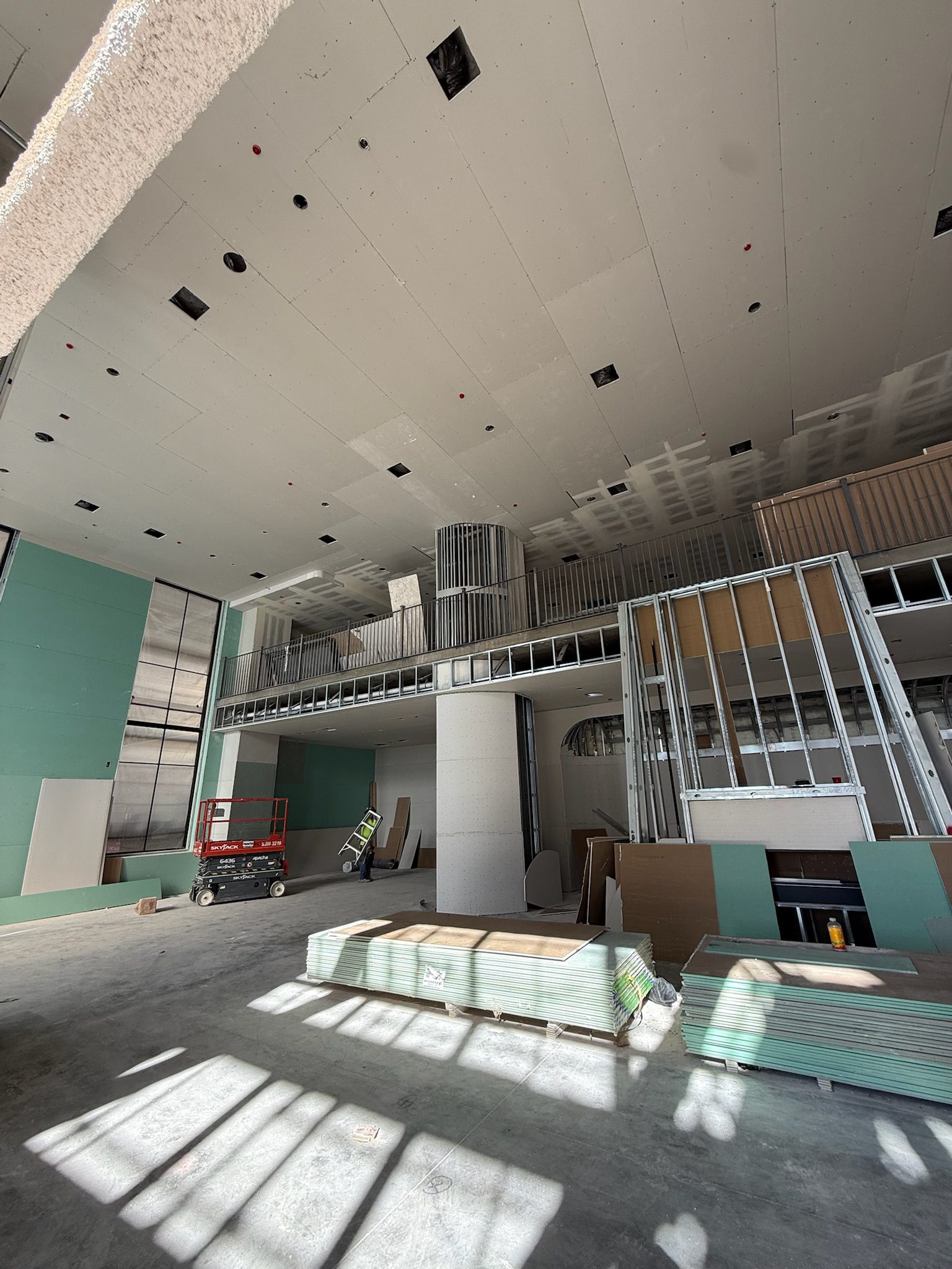 A large room under construction with unfinished walls and ceiling, exposed metal framing, drywall sheets, a red lift, and construction materials scattered on the concrete floor. Sunlight streams in through tall windows.