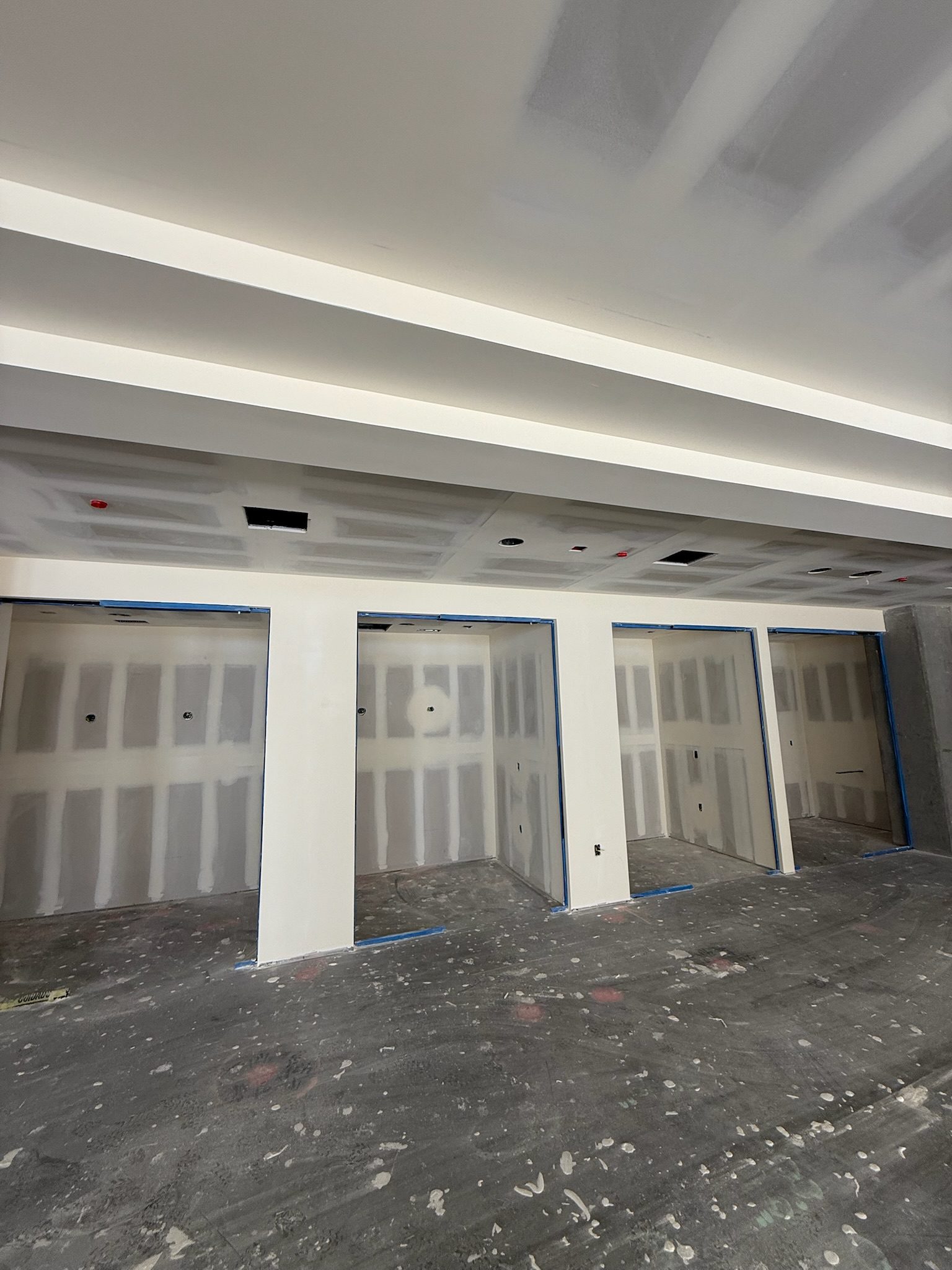 A room under construction with unfinished drywall, exposed studs, and several open wall sections. The ceiling and floor are unpainted, and blue painter’s tape lines some edges. Debris is scattered on the concrete floor.