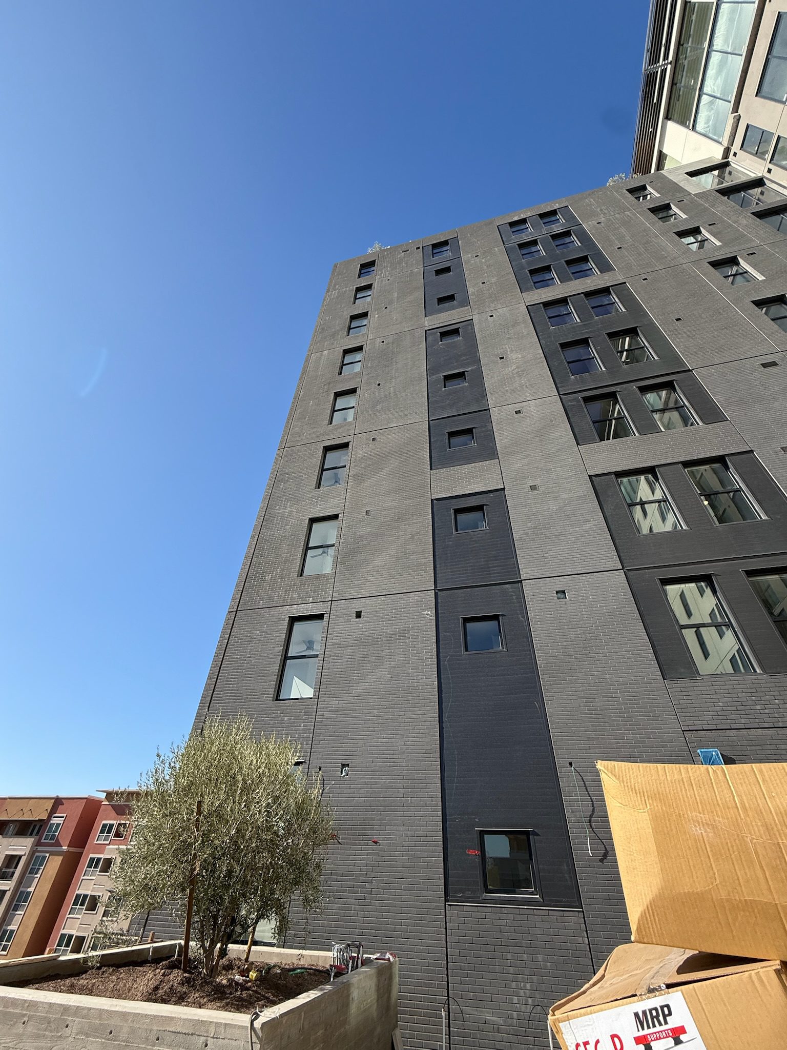 A tall, dark gray modern building with multiple windows is seen from a low angle against a clear blue sky. In the foreground, there’s a young tree in a planter and a cardboard box. Other buildings are visible to the left.