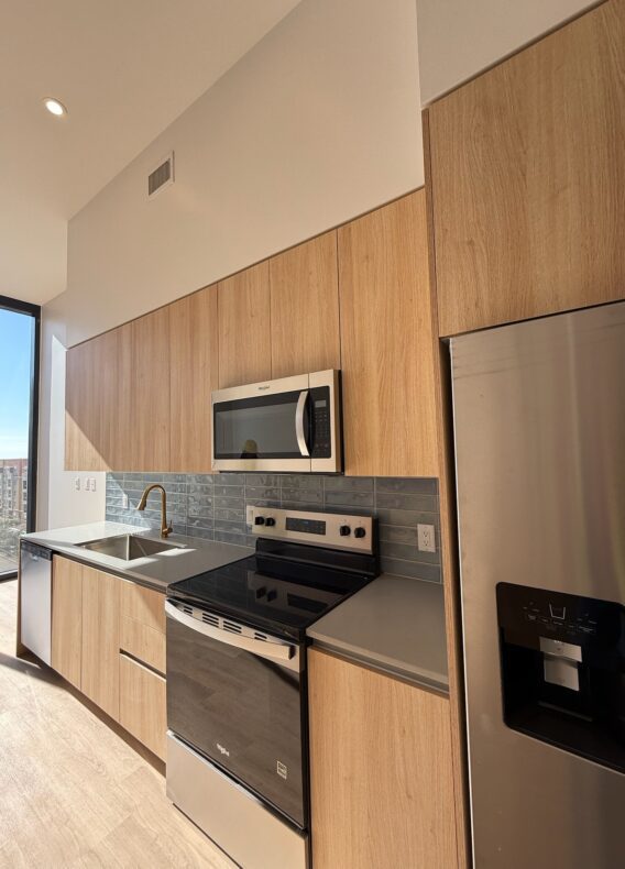 Modern kitchen with light wood cabinets, stainless steel appliances including a refrigerator, oven, microwave, and a sink under a window with sunlight streaming in. Gray tile backsplash and neutral countertops.