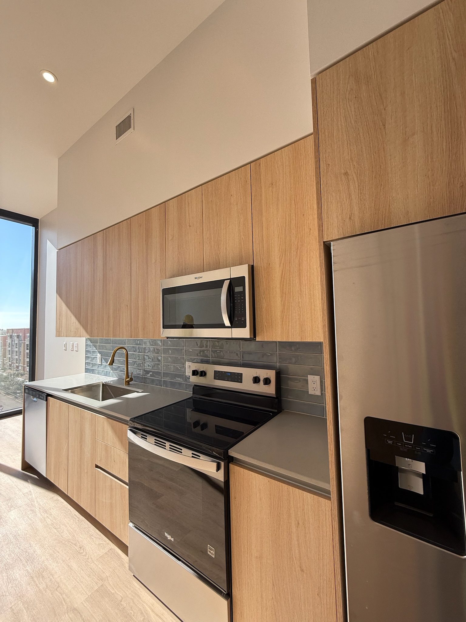 Modern kitchen with light wood cabinets, stainless steel appliances including a refrigerator, oven, microwave, and a sink under a window with sunlight streaming in. Gray tile backsplash and neutral countertops.