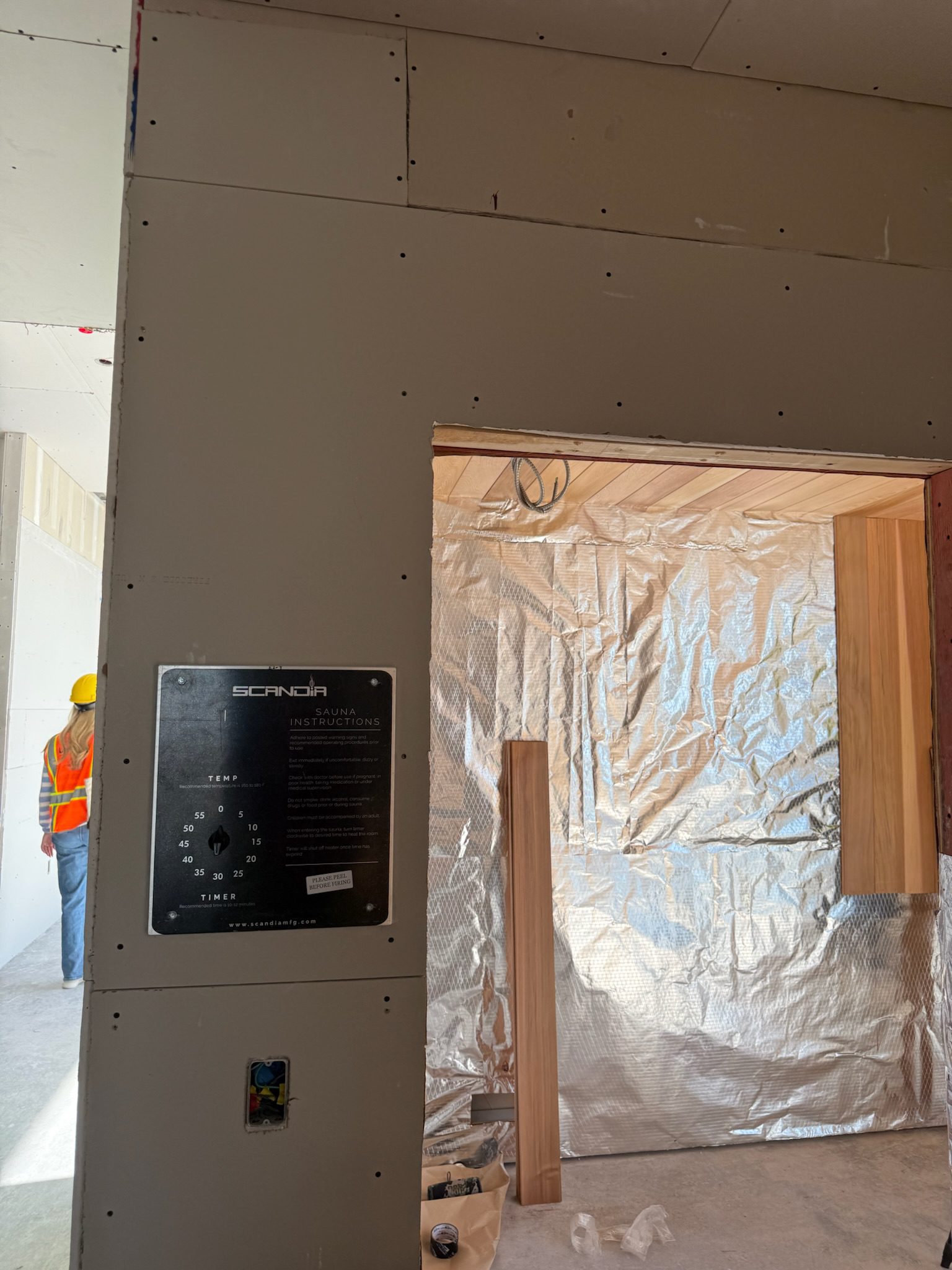 A construction site with a partially finished wall, exposed wiring, and a metal panel labeled SCANIA. A worker in a yellow hard hat and orange vest stands in the background, and foil insulation covers part of the wall.