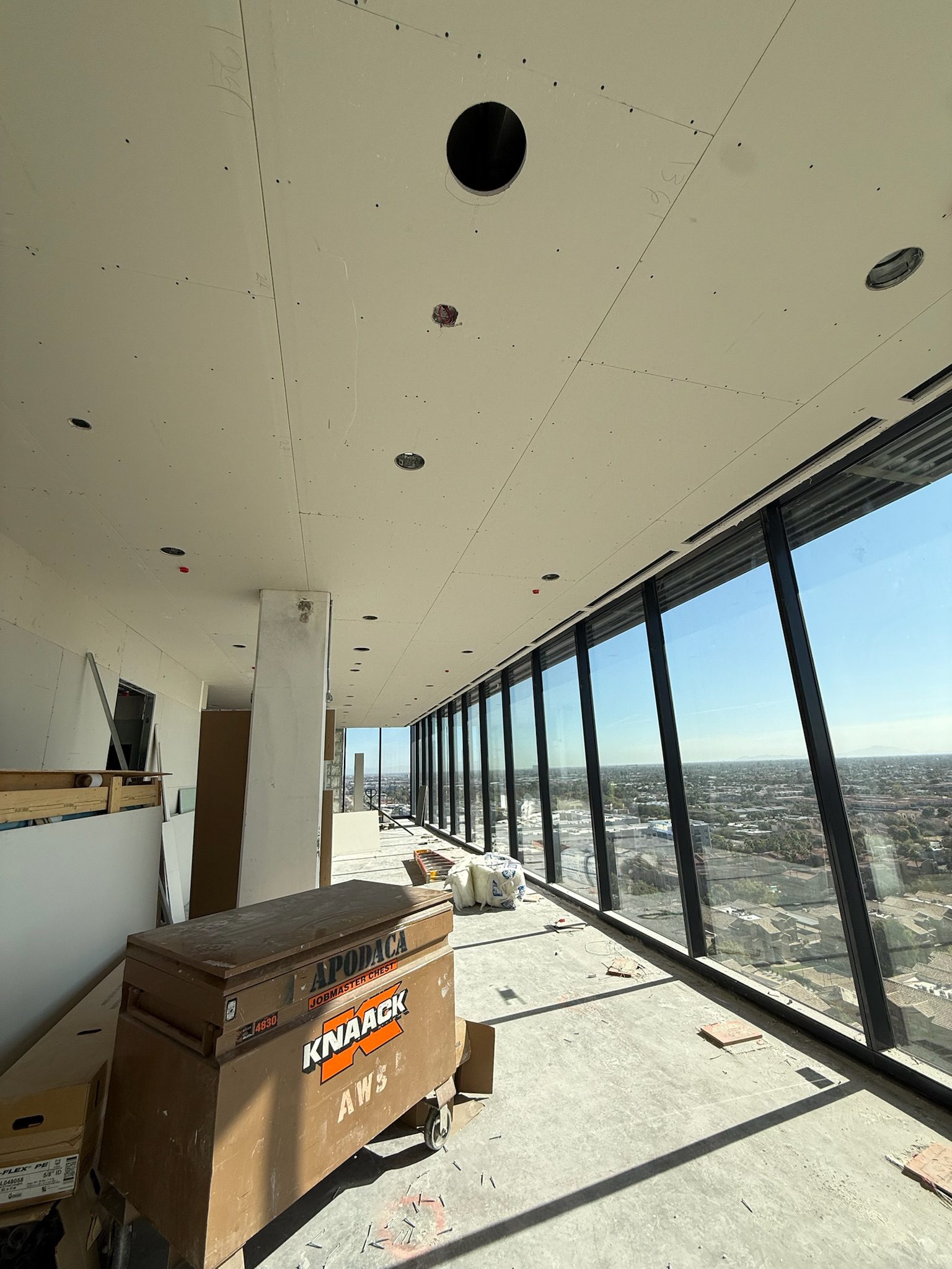 A high-rise building under construction with unfinished floors and ceiling, large floor-to-ceiling windows, and a cityscape view. Construction materials, boxes, and tools are scattered around the space.