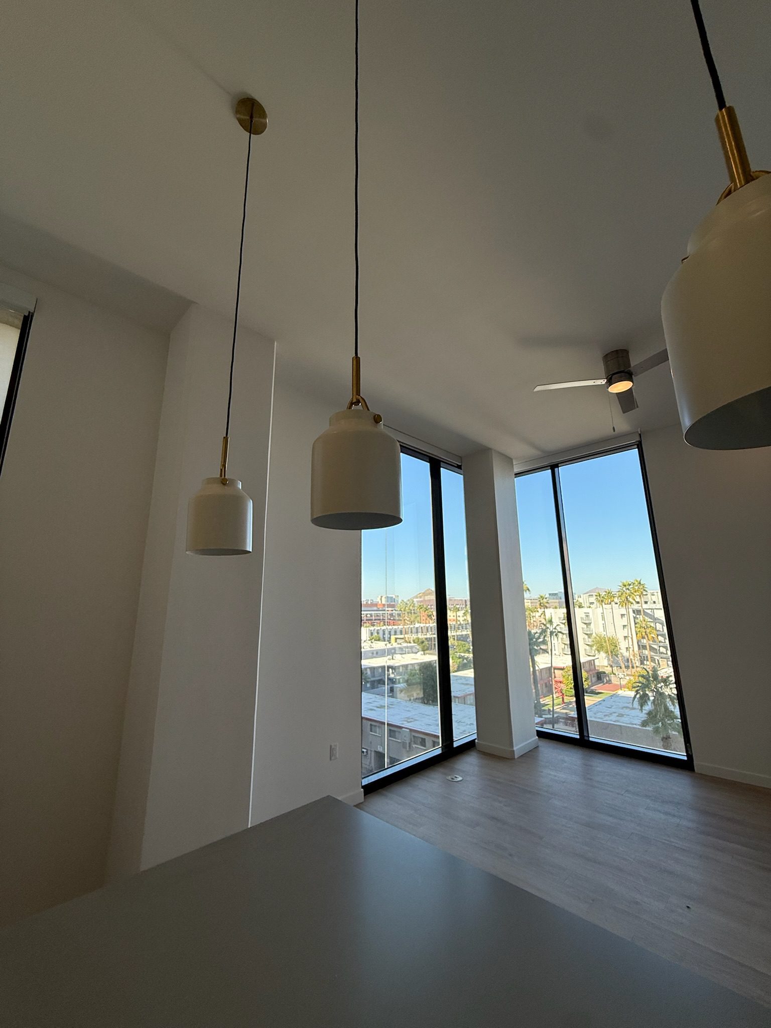 A modern, empty apartment with large floor-to-ceiling windows, wooden floors, and hanging pendant lights. Sunlight streams in, offering a clear view of city buildings and palm trees outside.