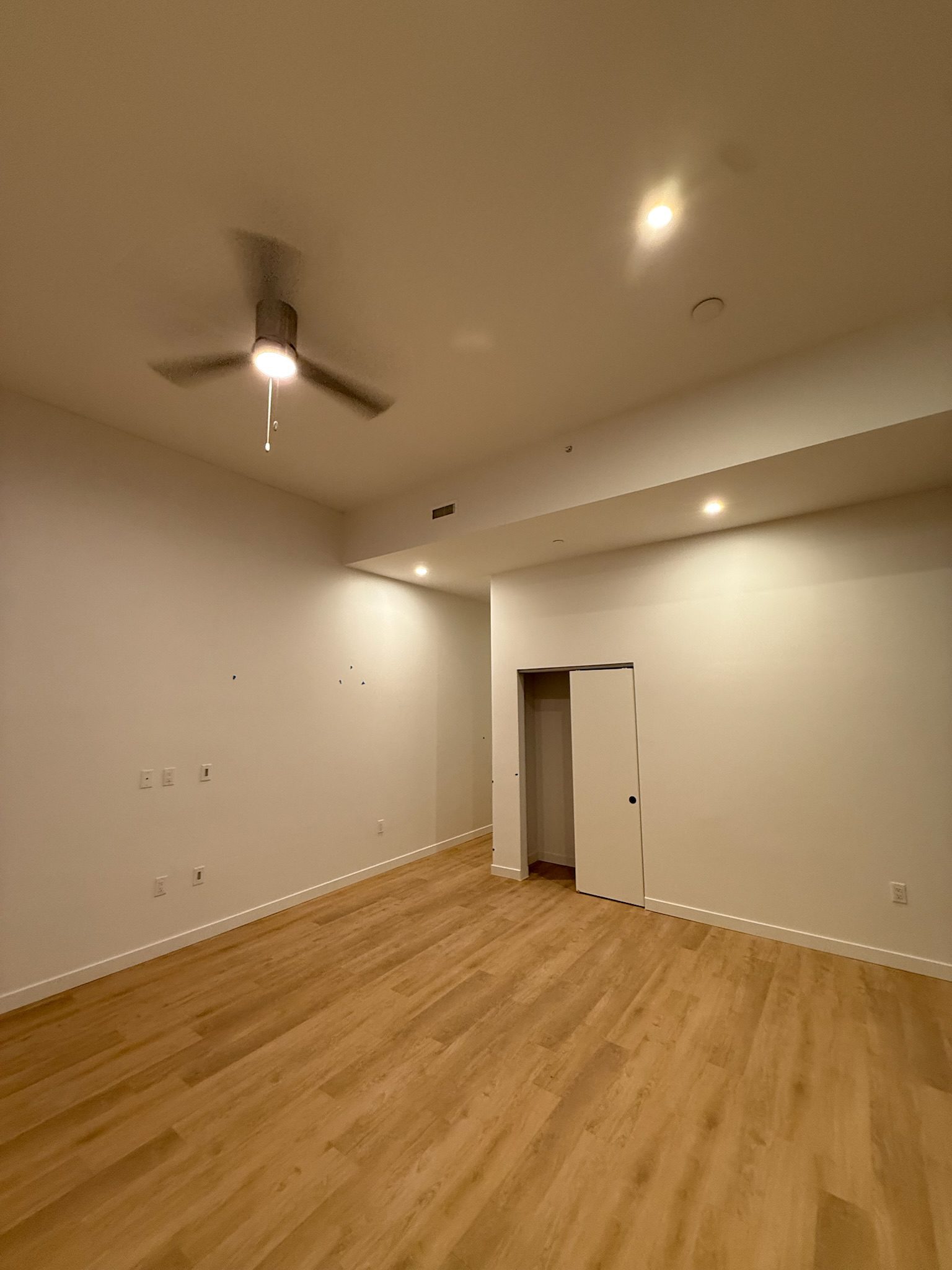 A spacious, empty room with light wood flooring, white walls, recessed lighting, a ceiling fan, and a closet with a sliding door. The overall lighting is warm and the ceiling is high.