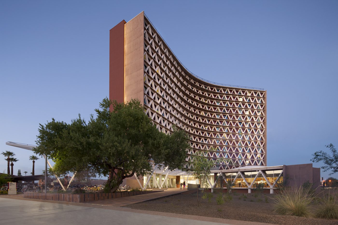 A tall, curved, modern building with a geometric patterned facade stands at dusk, surrounded by trees and desert landscaping, with lights glowing inside and people near the entrance.