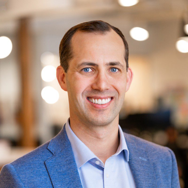 A man with short brown hair, wearing a blue blazer and light blue collared shirt, smiles at the camera in a softly lit indoor setting with blurred lights and background.