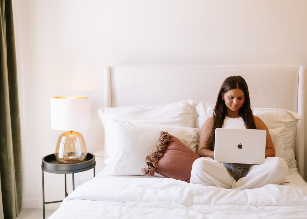A freshman woman sits cross-legged on a neatly made bed with white bedding, using a laptop. A lamp on a round black side table is beside her. The room appears bright and minimalistic.