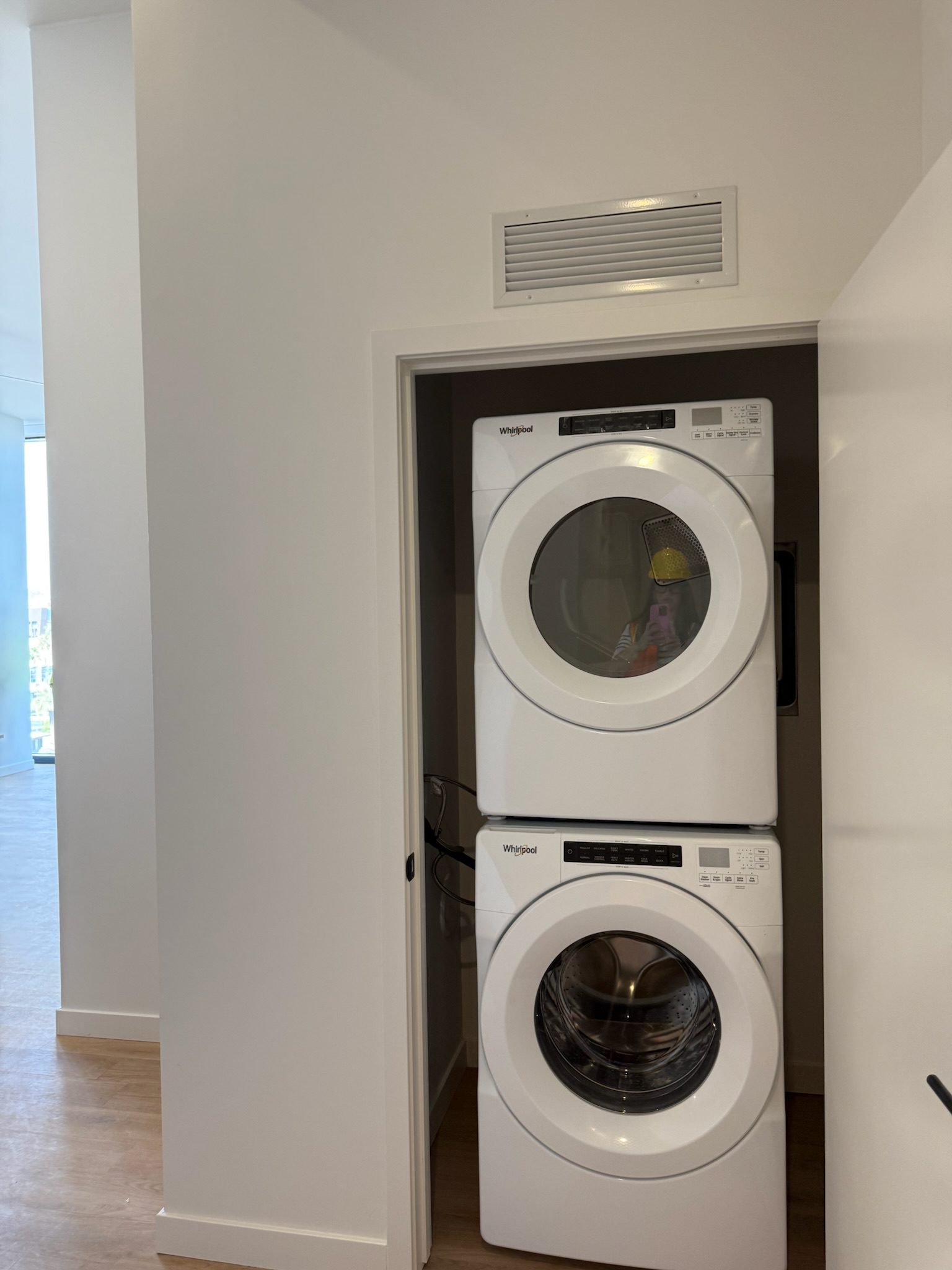 A stacked Whirlpool washer and dryer sit in a small, white laundry closet with a vent above—part of a recent construction update. The closet door is partially open, and both machines feature digital control panels.