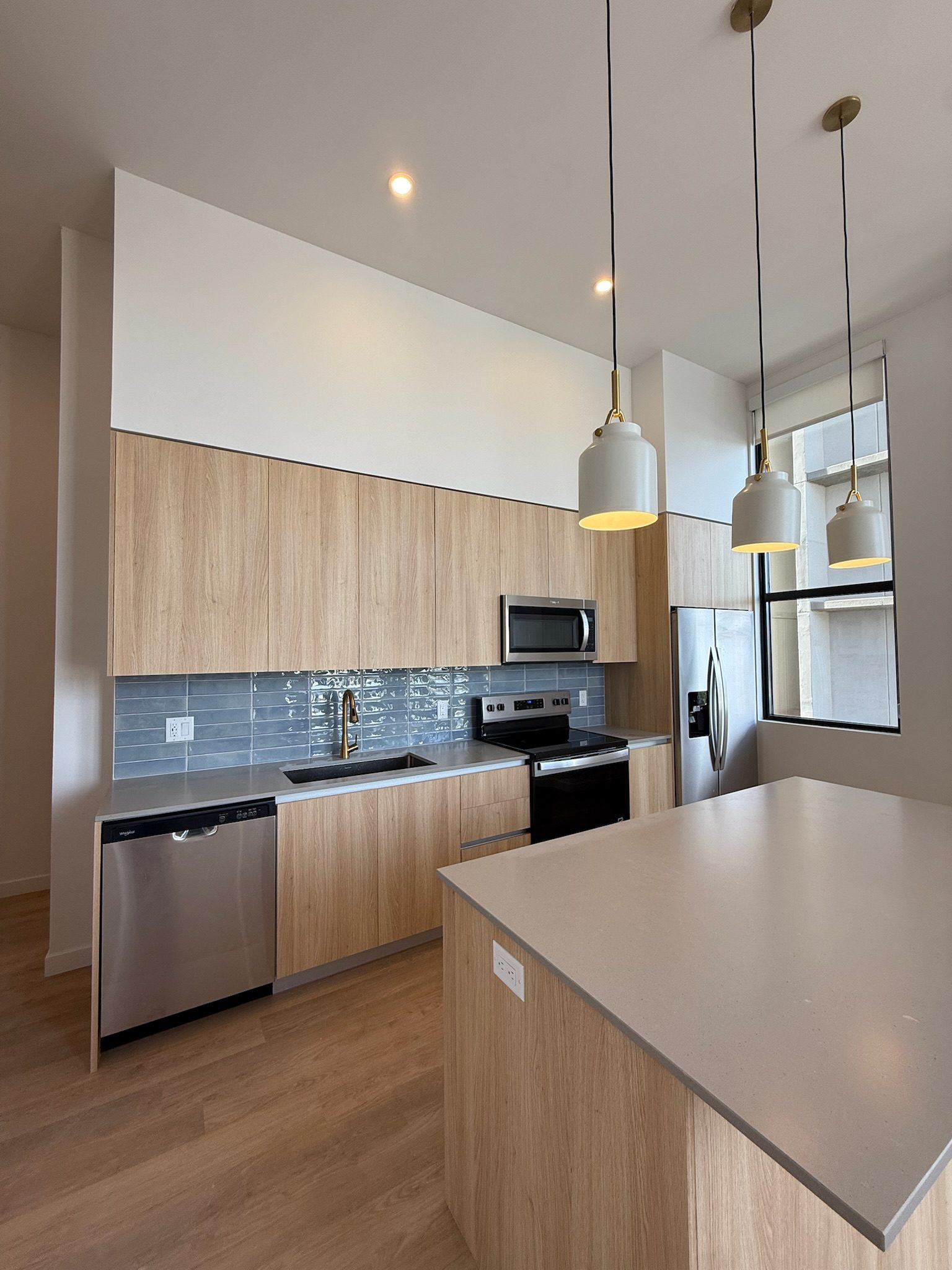 Modern kitchen with light wood cabinets, stainless steel appliances, a large island with a light gray countertop, pendant lights, and a window providing natural light—perfect for your next construction update.