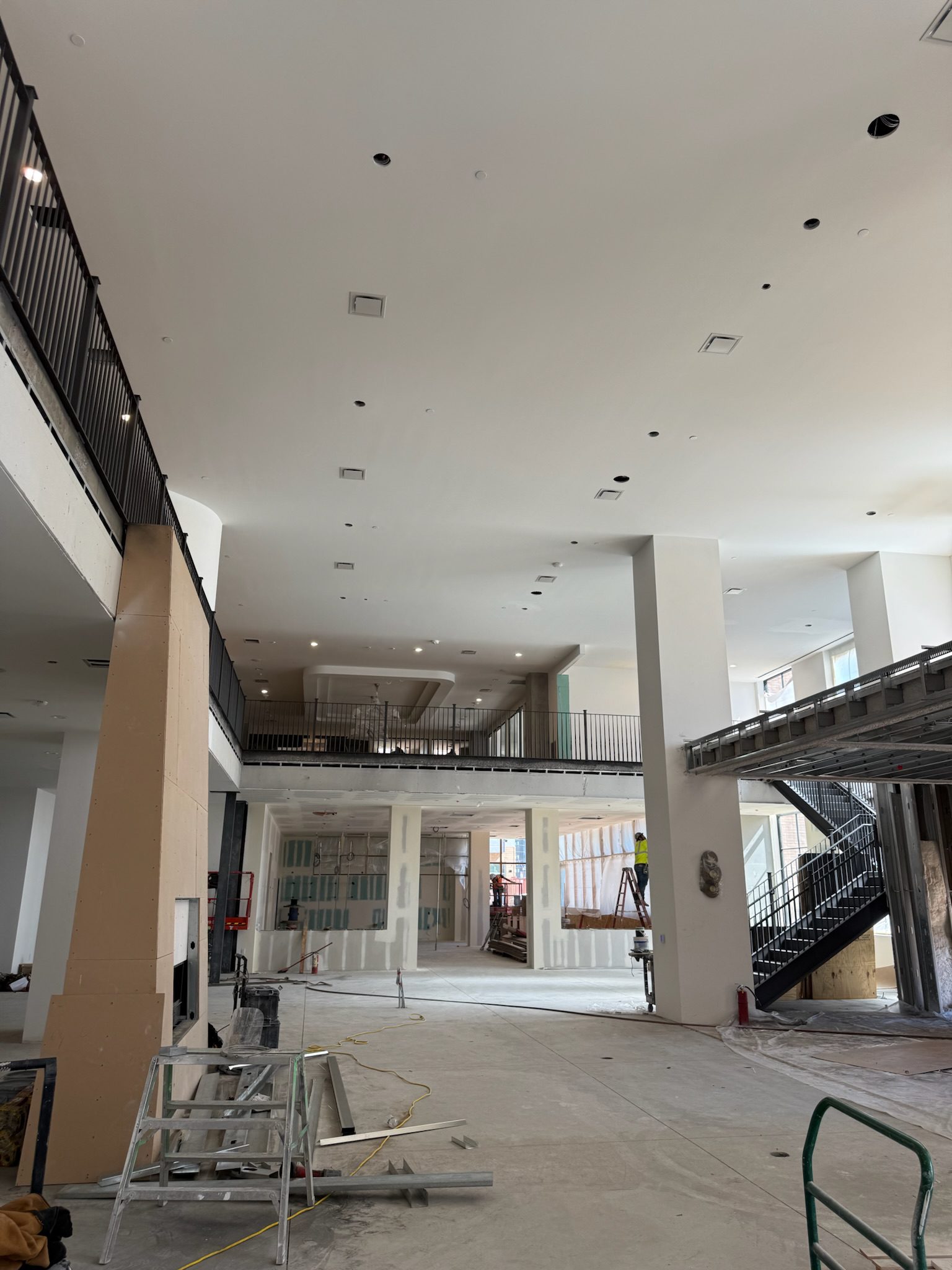 Construction update: A large indoor space under construction with exposed drywall, unfinished floors, ladders, and materials. A staircase leads to a mezzanine with black railings, while large windows fill the area with natural light.