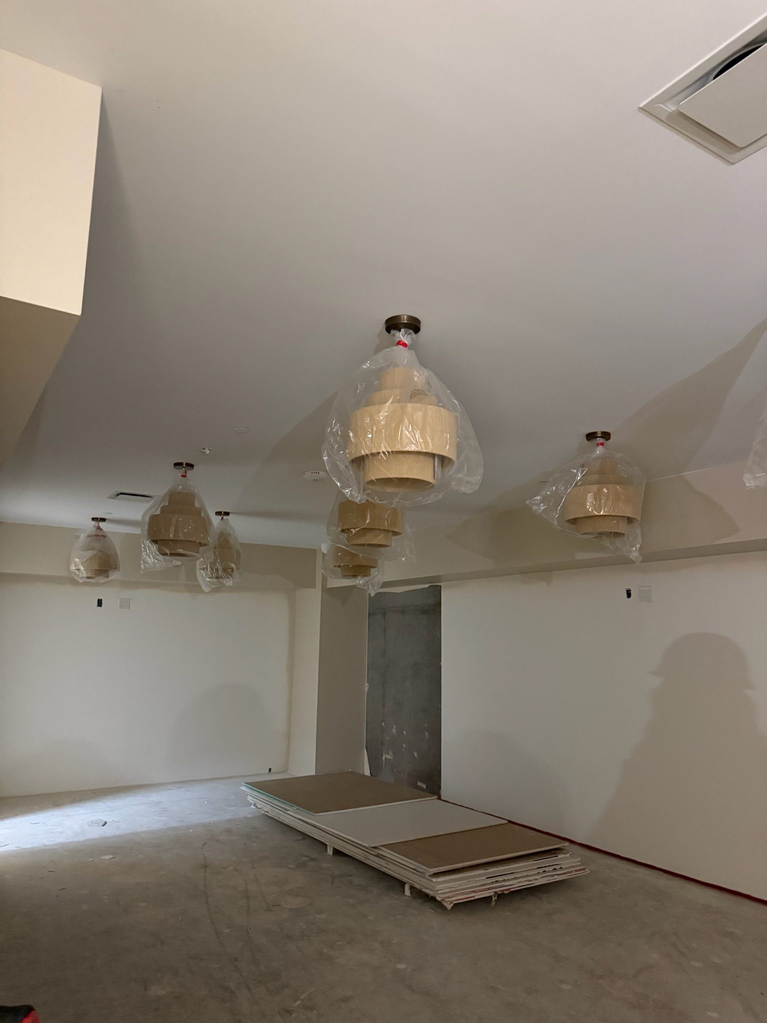 Several light fixtures wrapped in plastic hang from the ceiling in an unfinished room, offering a clear construction update with bare walls and stacked boards scattered on the floor.