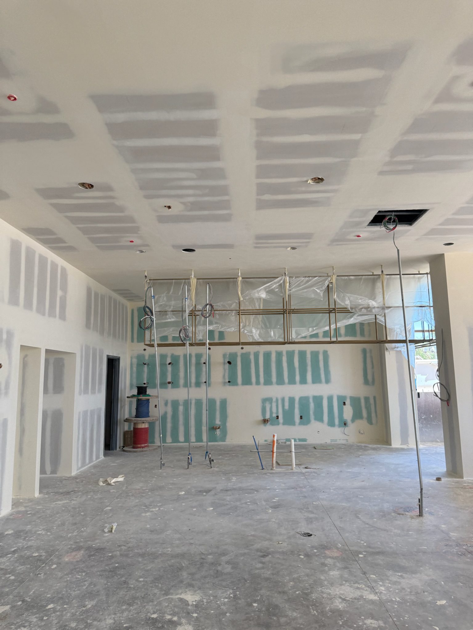 An unfinished room under construction with drywall installed, exposed wiring hanging from the ceiling, unpainted walls, and construction materials on the floor. Plastic sheeting covers part of the back wall in this latest construction update.