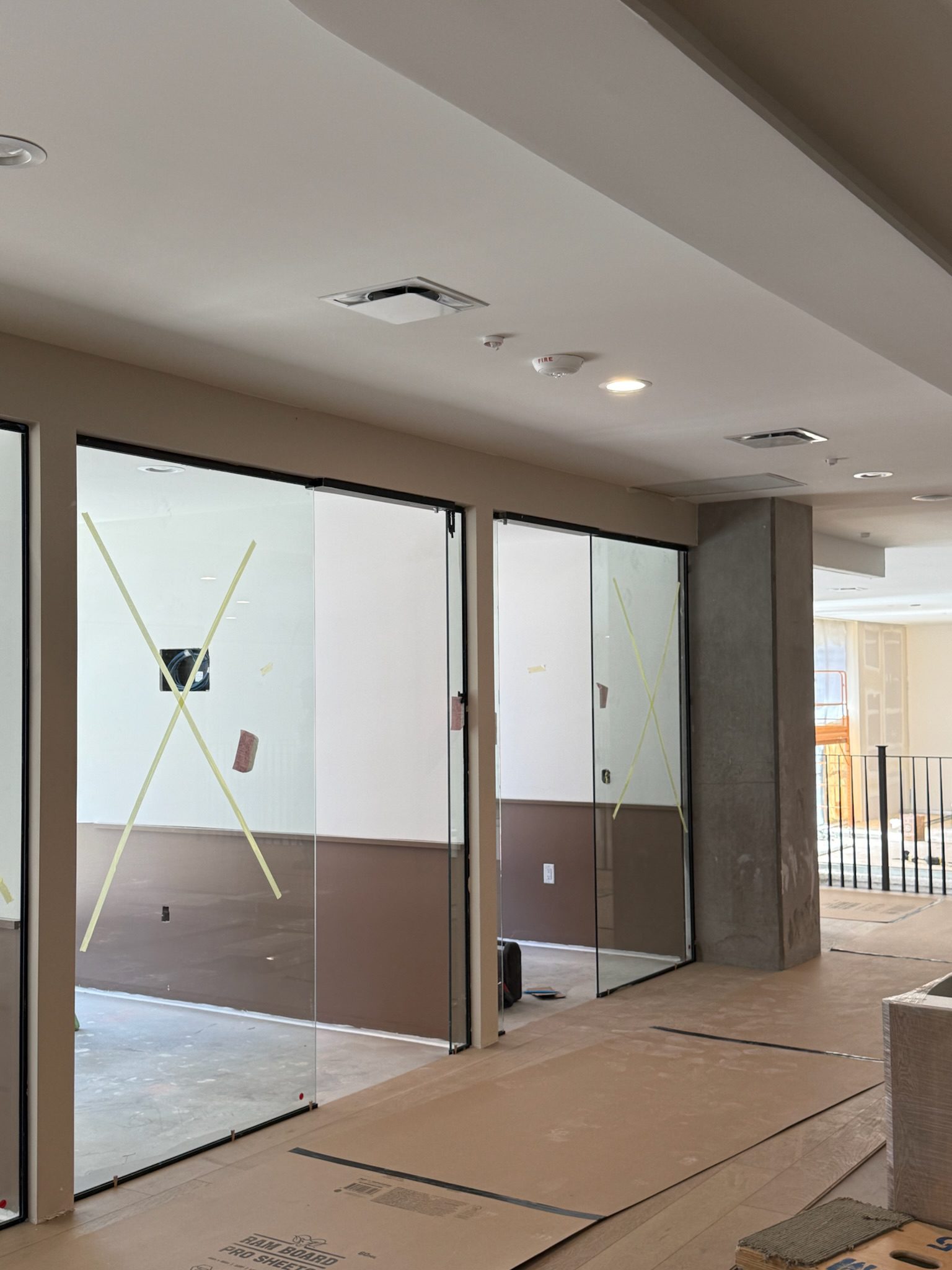 A modern interior under construction update features large glass walls marked by yellow tape in X shapes, brown trim, exposed concrete columns, and protective floor coverings. Natural light enters from the right side.