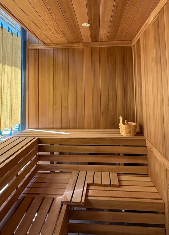 A modern wooden sauna room with tiered benches, a wooden bucket with a ladle, and sunlight streaming in through large windows covered by partially drawn curtains showcases the latest construction update.