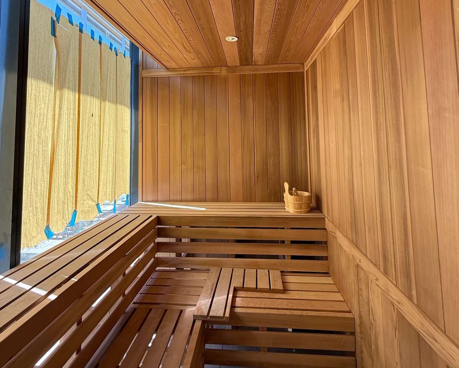 A modern wooden sauna room with tiered benches, a wooden bucket with a ladle, and sunlight streaming in through large windows covered by partially drawn curtains showcases the latest construction update.