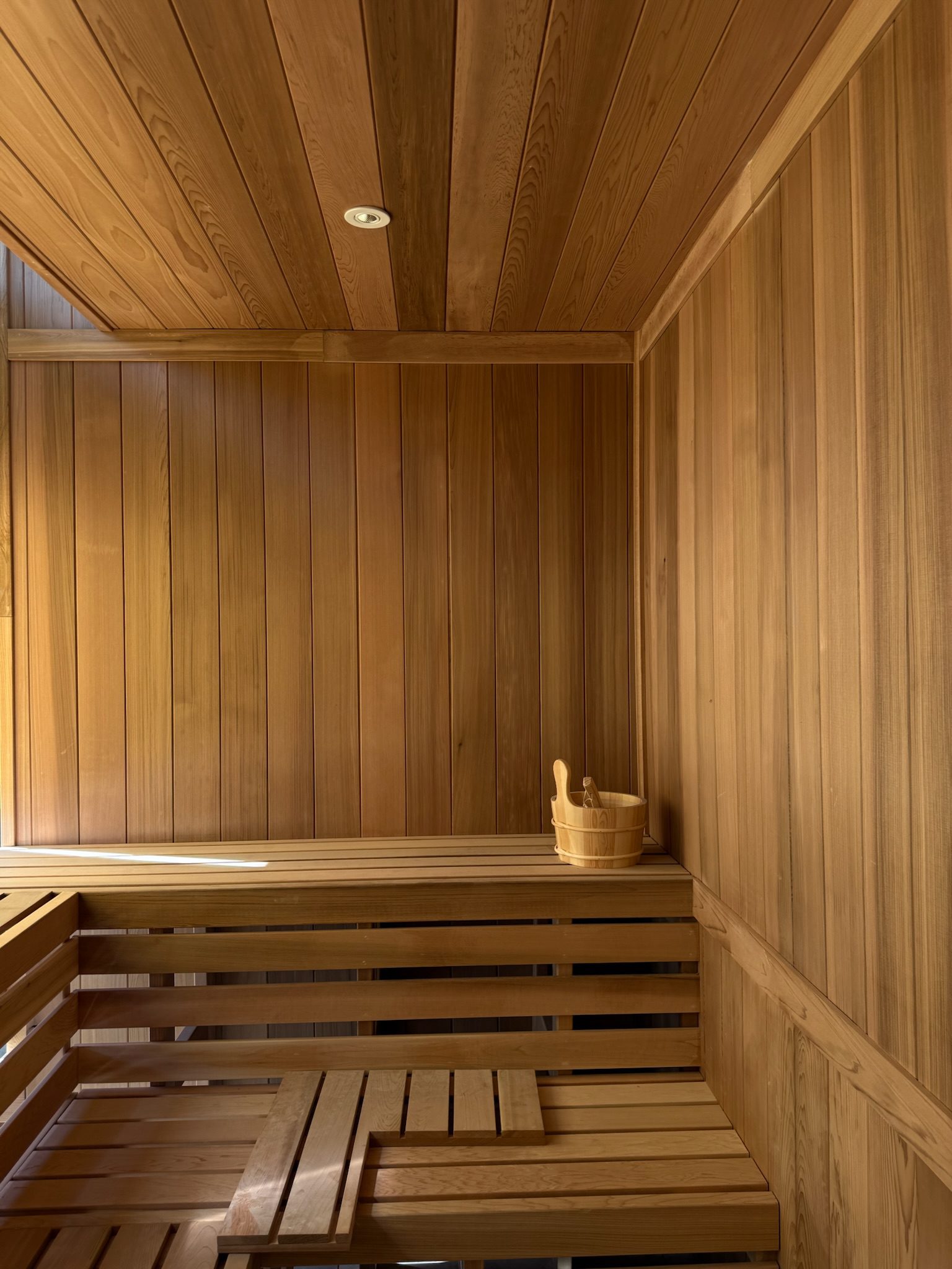 A wooden sauna interior with benches and a bucket with a ladle in the corner. Warm light highlights the smooth, natural wood panels covering the walls, benches, and ceiling—a stunning construction update for any relaxation space.