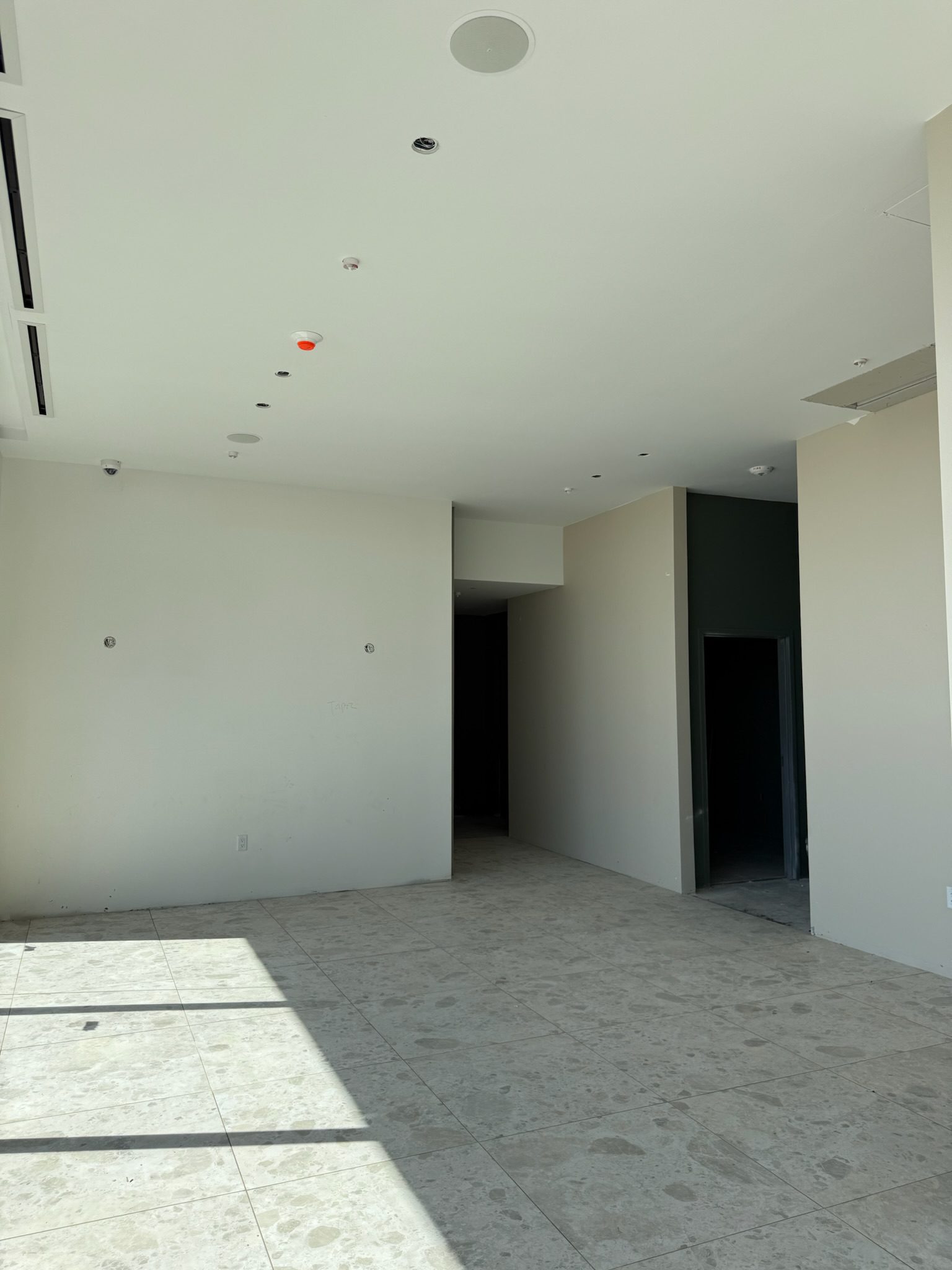 A mostly empty room with light-colored walls and tiled floors, featuring a doorway leading to a dark hallway. Sunlight shines in from the left, highlighting recent construction updates with bright patches on the floor.