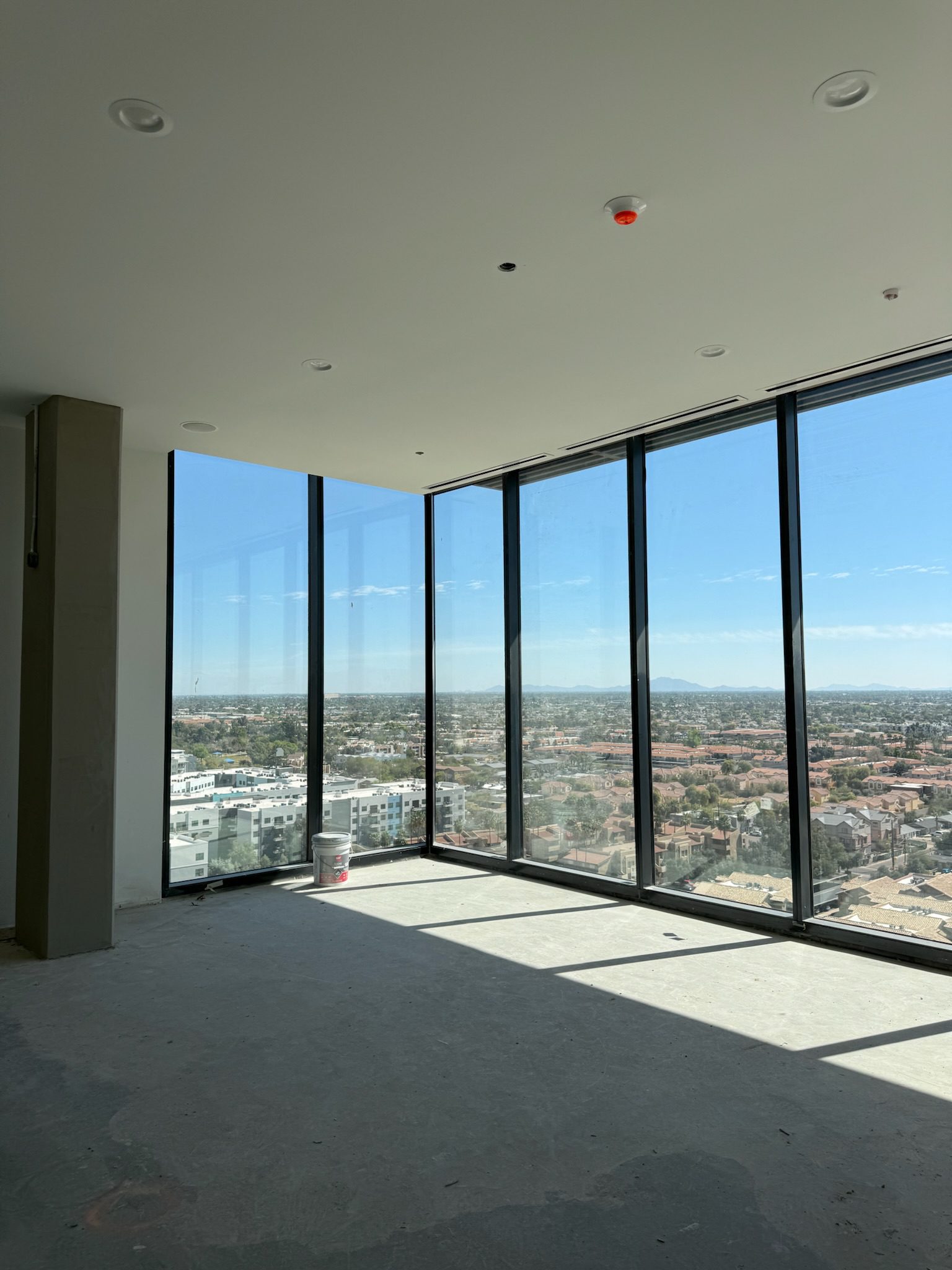 A mostly empty room with large floor-to-ceiling windows offers a clear view of the cityscape under a blue sky. Sunlight casts shadows on the unfinished concrete floor—a paint bucket near the windows hints at an ongoing construction update.