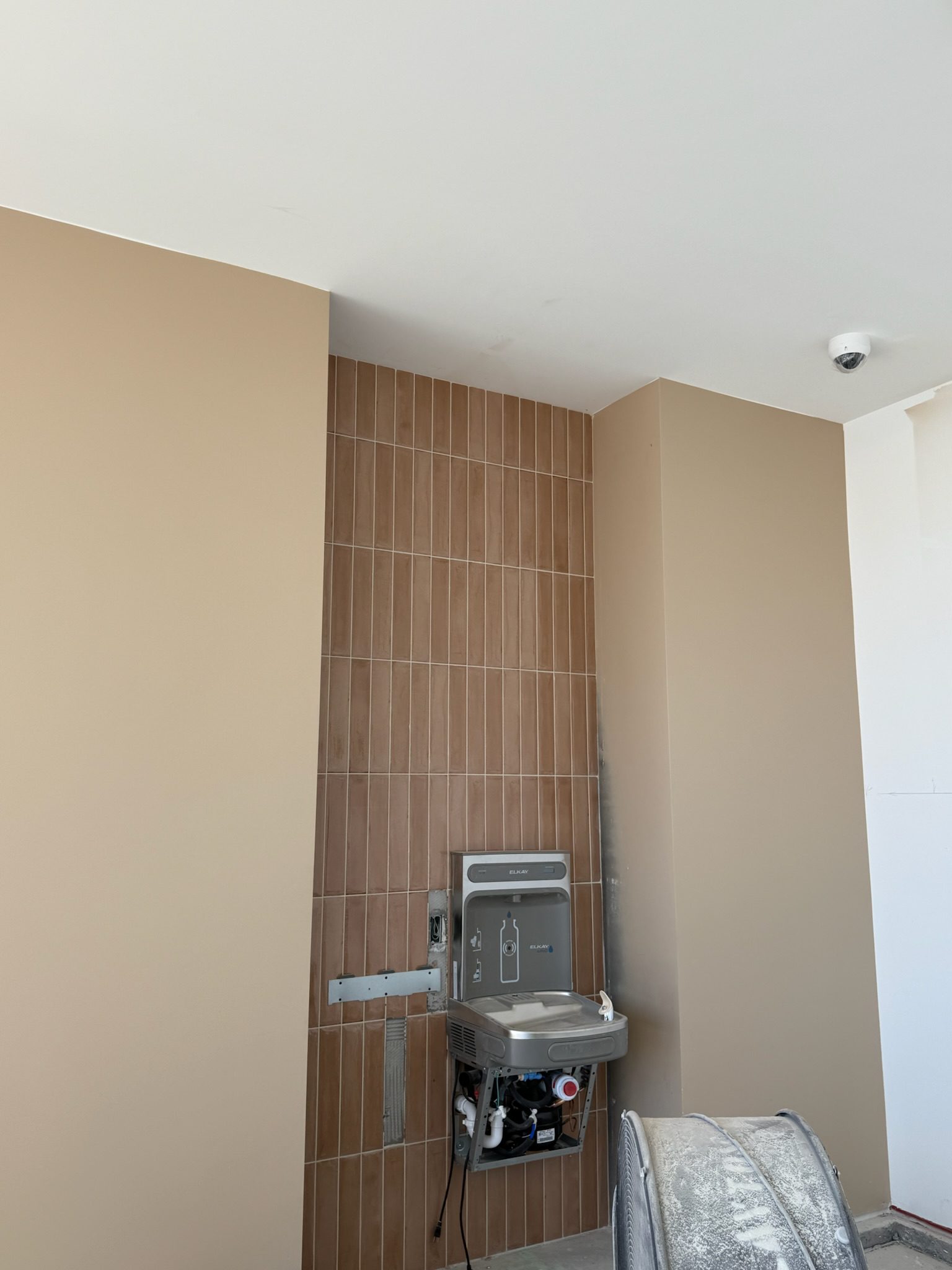 A stainless steel drinking fountain and bottle filling station are installed against a wall with vertical brown tiles, flanked by two beige painted columns. As part of the recent construction update, exposed pipes and a security camera are visible above.