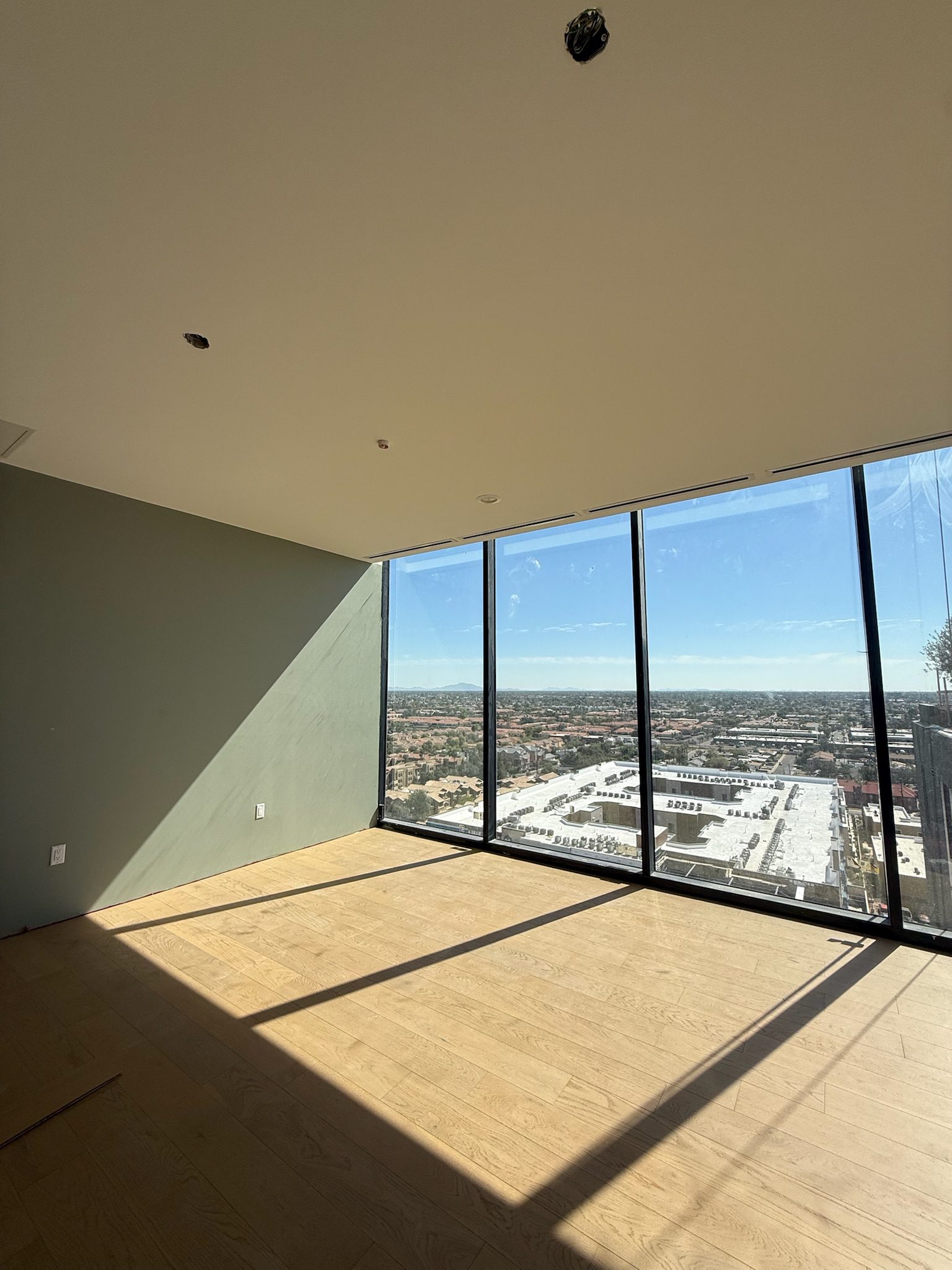 Sunlight streams into an empty room with light wooden floors and large floor-to-ceiling windows, showcasing a panoramic cityscape view under a clear blue sky—a stunning highlight in this latest construction update.