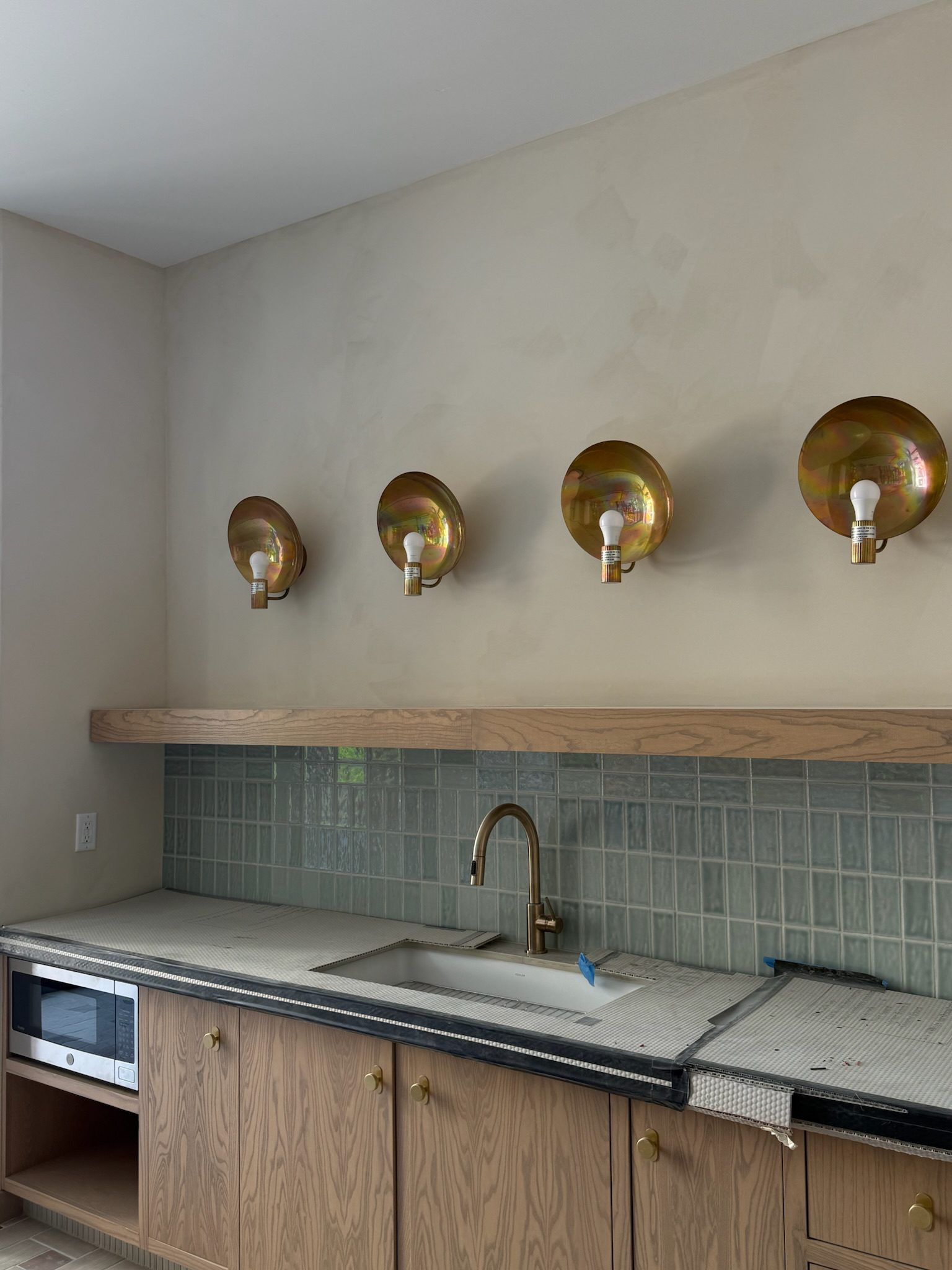 A modern kitchen under renovation gets a construction update: unfinished countertops, brass sink and faucet, a glass tile backsplash, and four brass wall sconces with exposed bulbs. A microwave is seamlessly built into the lower cabinets.