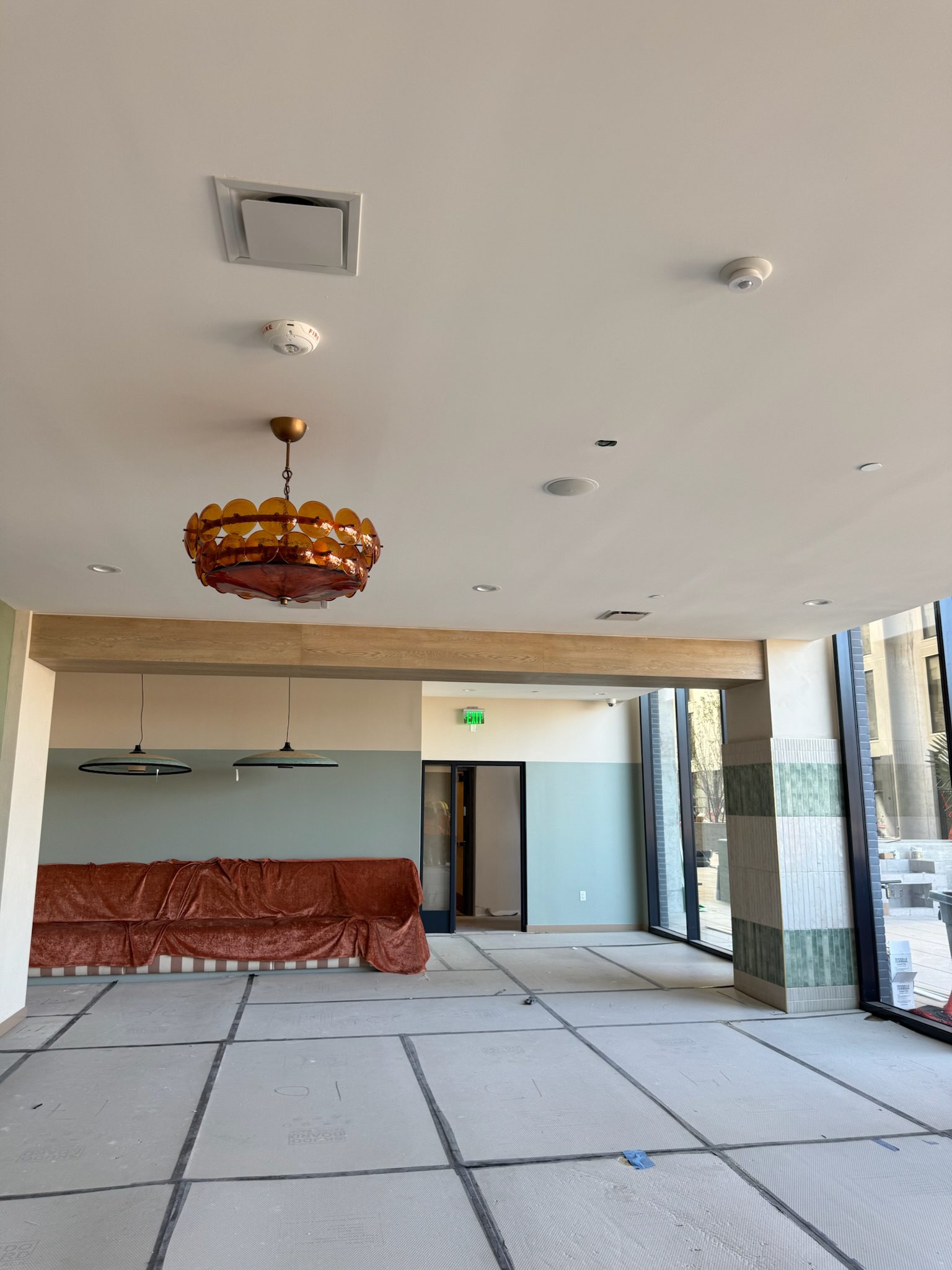 A spacious, unfinished room with covered furniture, large windows, a retro-style amber ceiling light, and protective floor coverings—a snapshot of a construction update in progress.