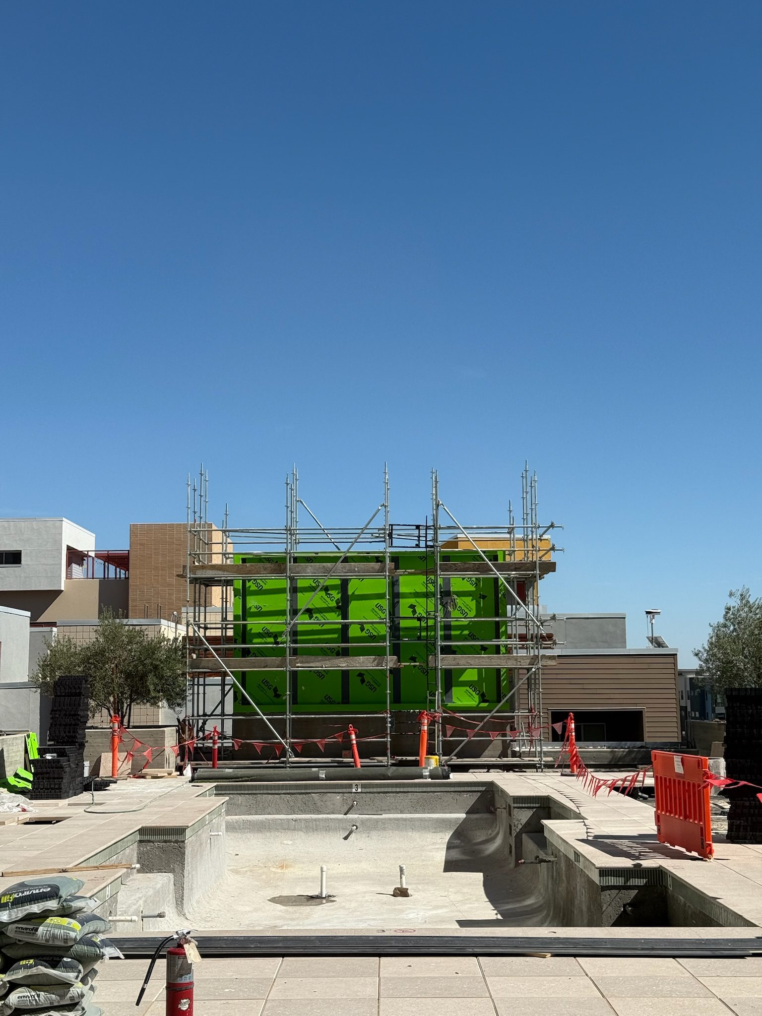 A construction update: Scaffolding surrounds a building with a bright green wall. In the foreground, an empty pool, construction materials, and orange safety barriers sit under a clear blue sky.
