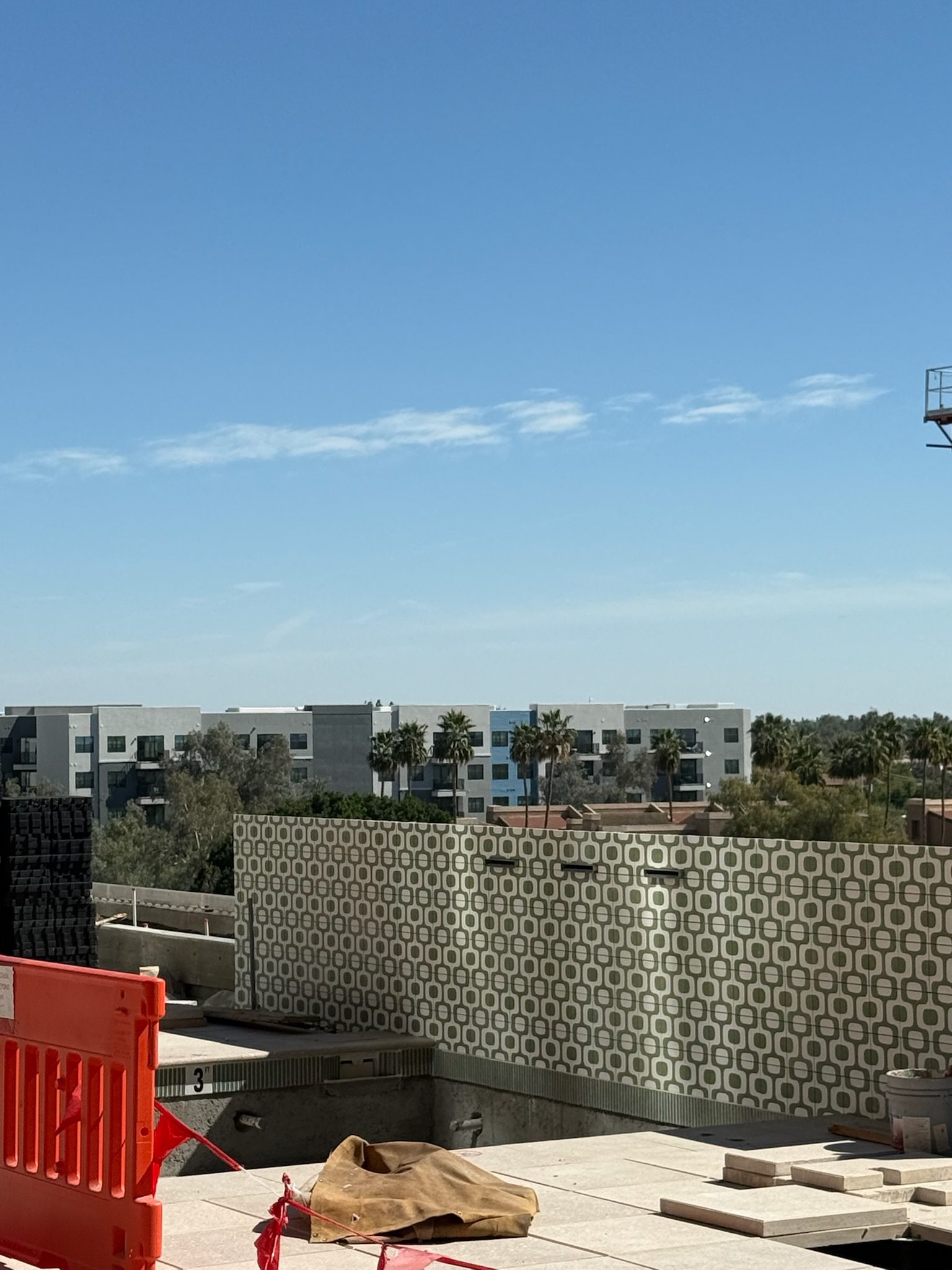 A rooftop construction area with an orange safety barrier, scattered materials, and a patterned wall. This construction update also shows modern apartment buildings, palm trees, and a clear blue sky in the background.