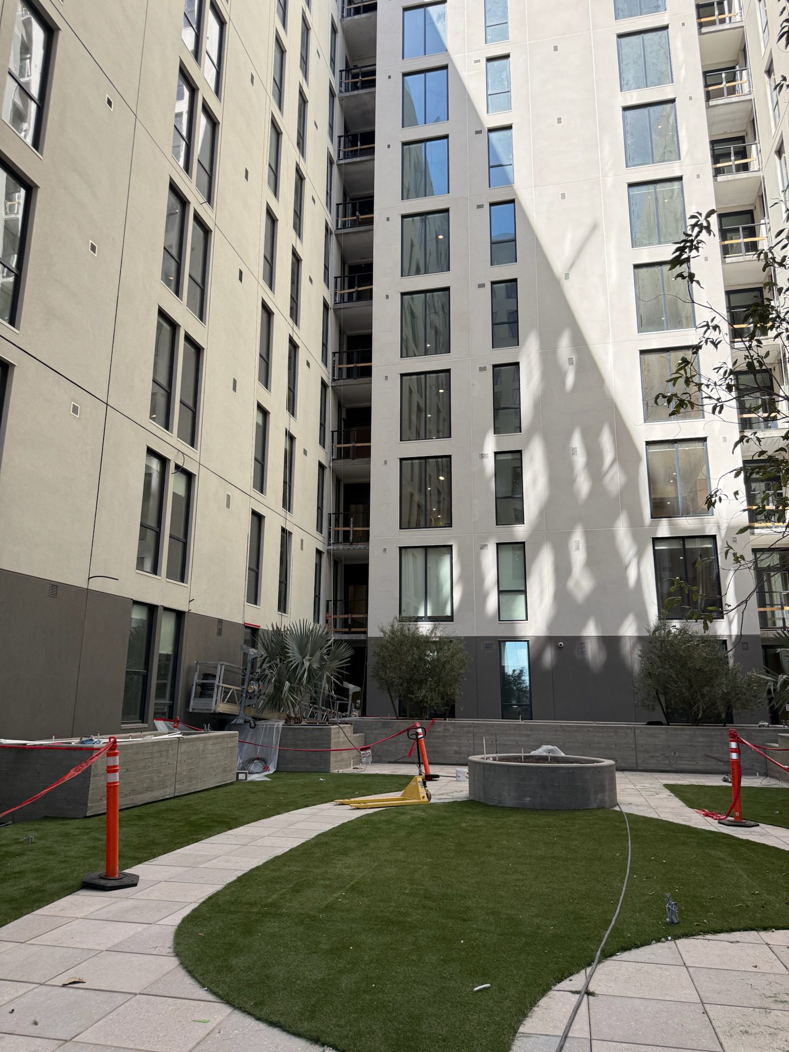 A modern apartment courtyard with artificial grass, a small circular fountain, and surrounding tall beige buildings. Caution tape and cones block off parts of the walkway, providing a clear construction update for residents.
