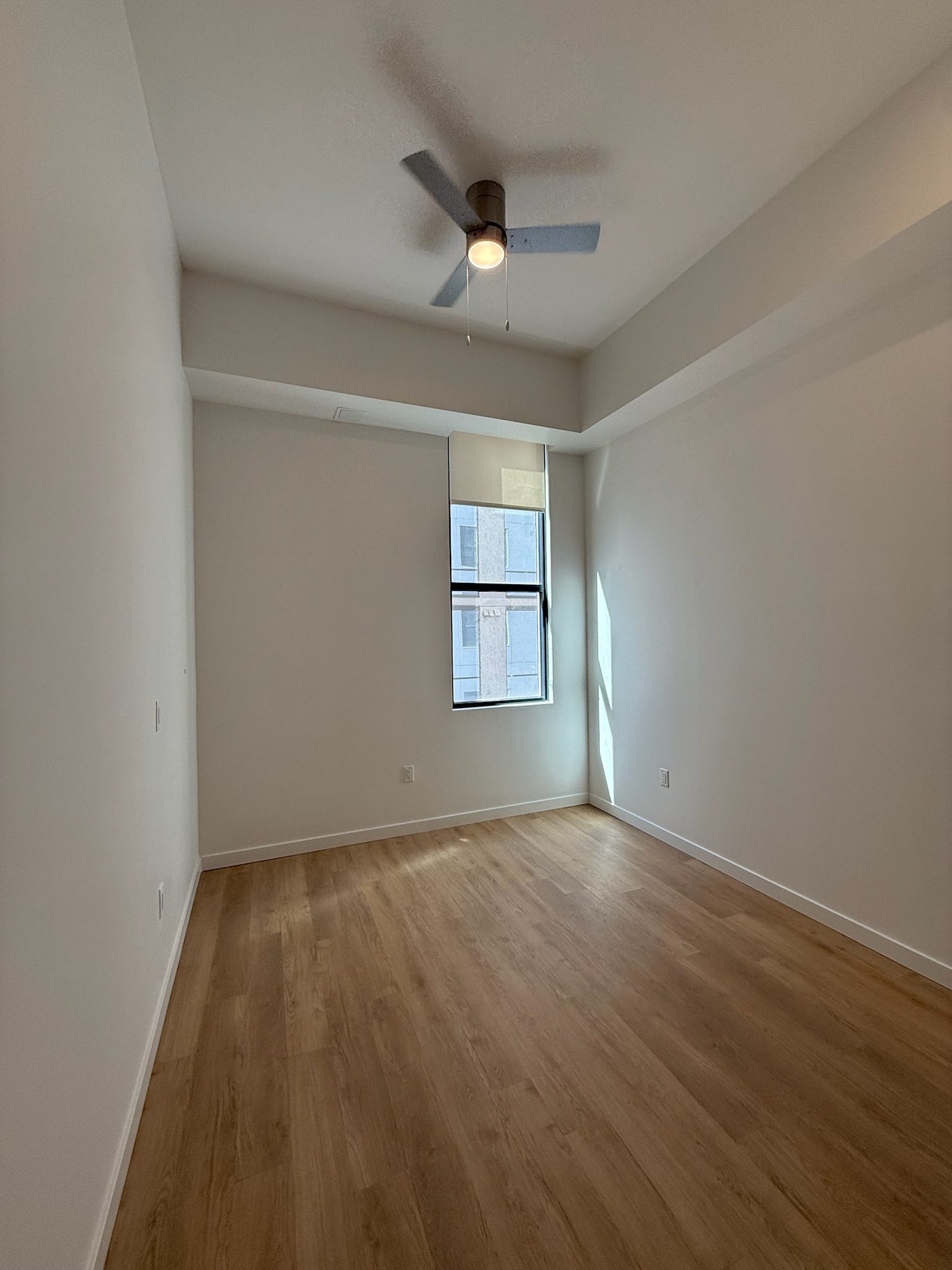 A bright, empty room with light wood flooring, white walls, a ceiling fan with light, and a window letting in natural daylight provides a fresh look in this latest construction update.