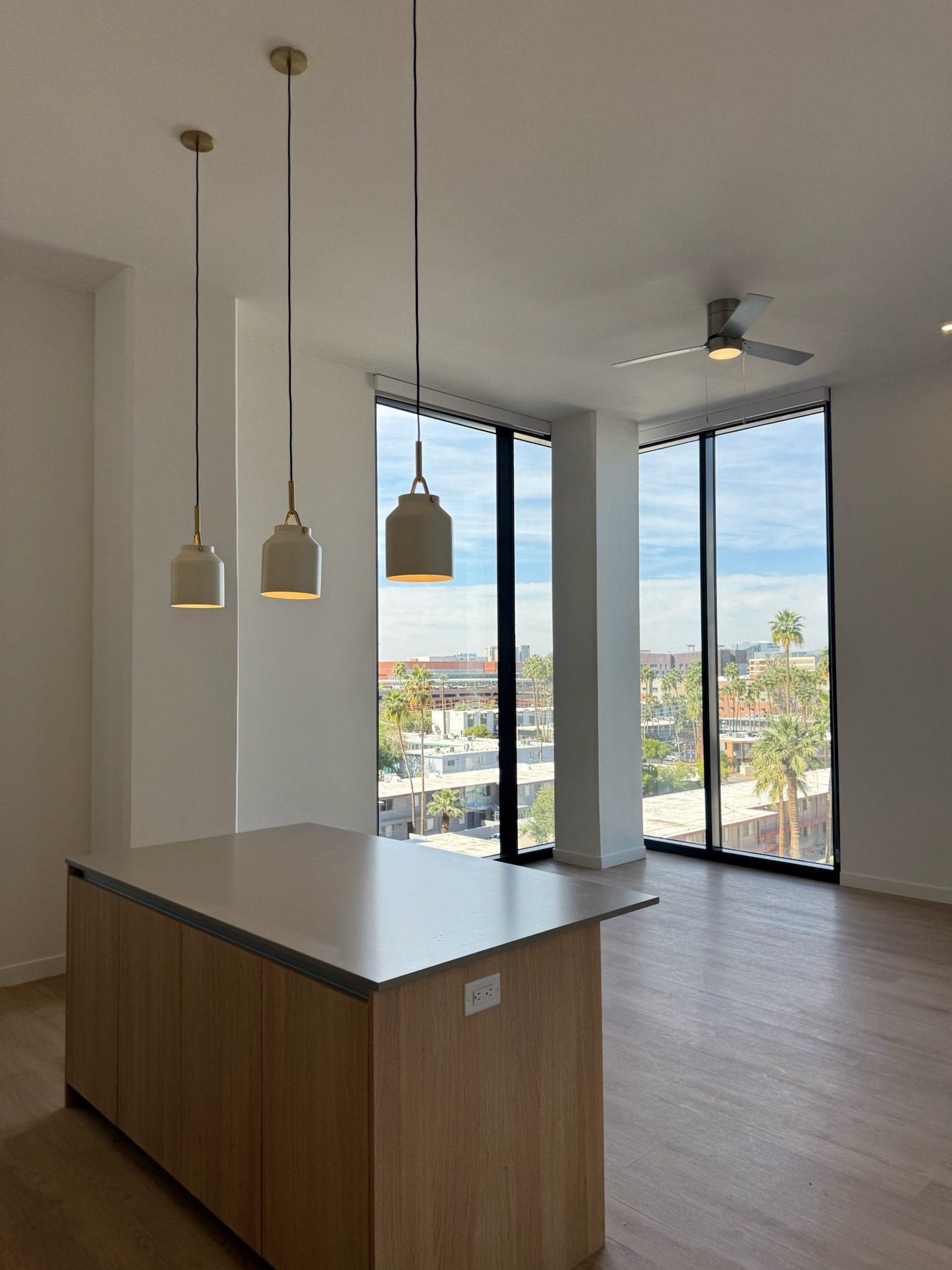Modern, unfurnished apartment with light wood floors, an island with pendant lights, large floor-to-ceiling windows, and a ceiling fan, overlooking a cityscape with palm trees—perfect for those following the latest construction update.