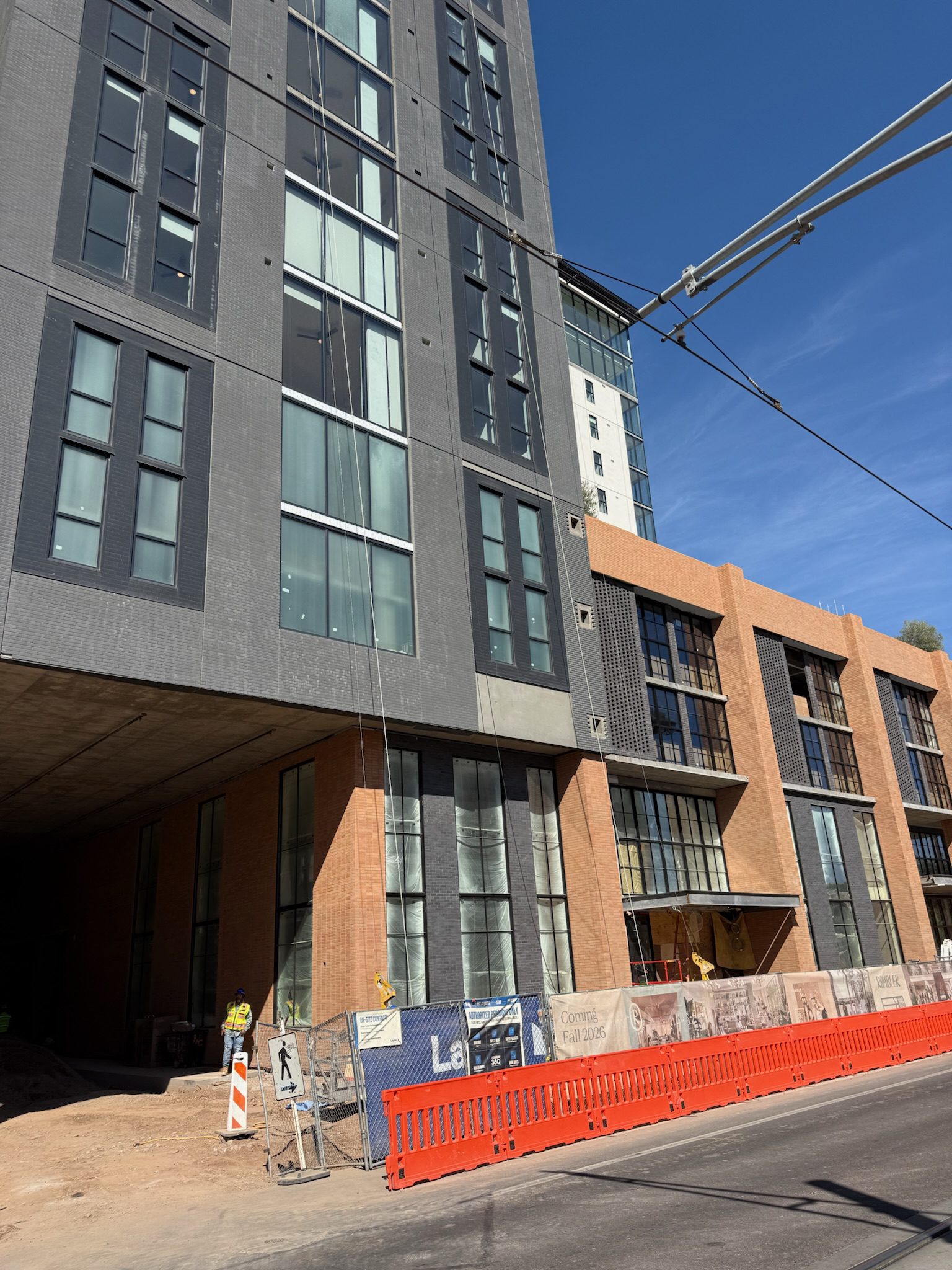 A modern multi-story building under construction with gray and brick exteriors. This construction update shows workers in safety gear near orange barriers along the sidewalk on a sunny day.