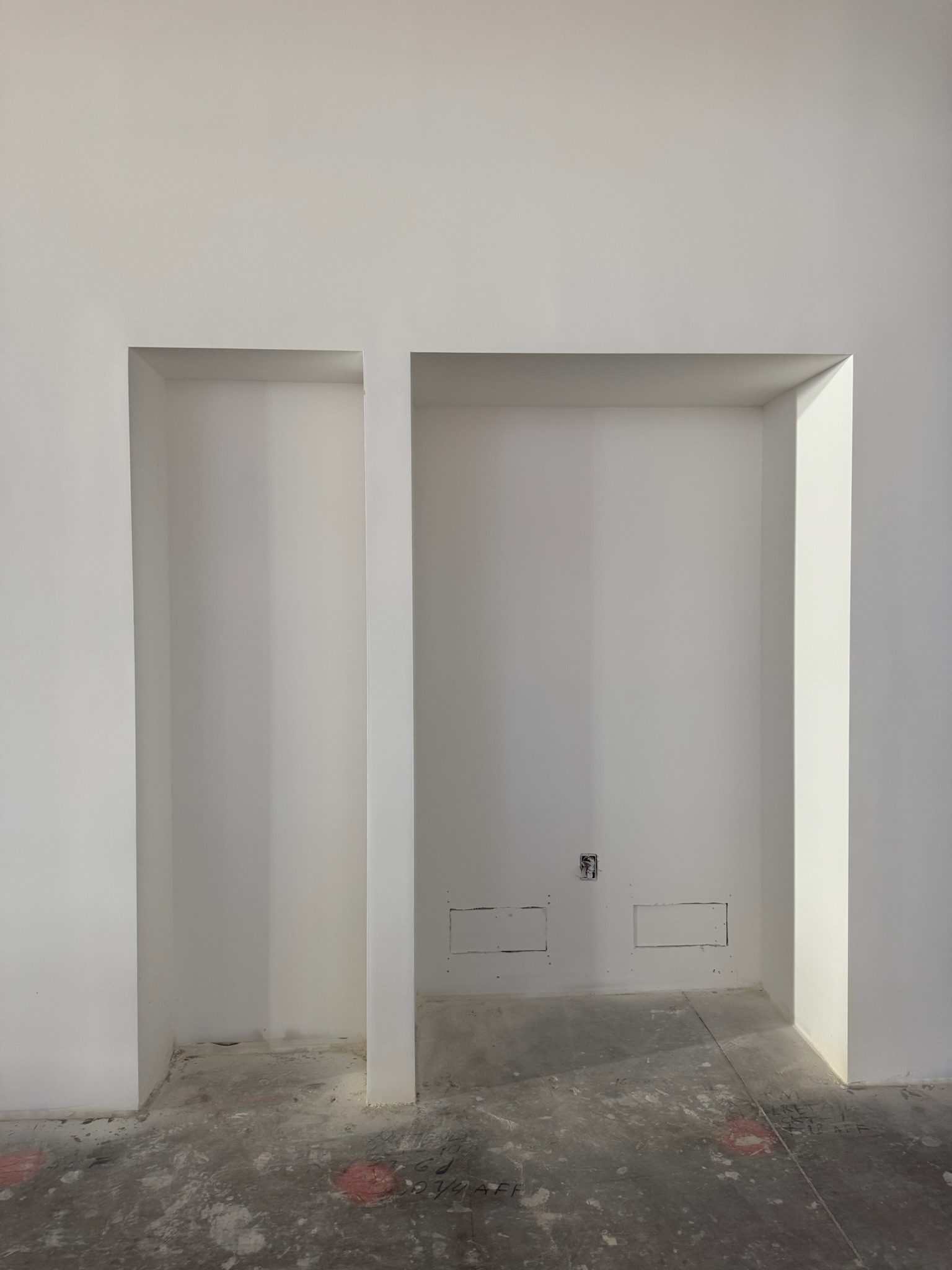 Construction update: A white wall with two recessed rectangular niches; the floor is unfinished concrete, and there are two small utility access panels and an electrical outlet in the right niche.