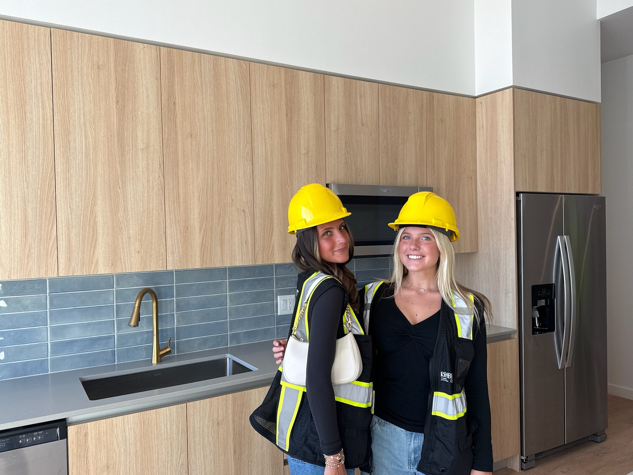 Two women wearing yellow construction helmets and safety vests smile during a hard hat tour in a modern kitchen with light wood cabinets, a gray tile backsplash, and stainless steel appliances.