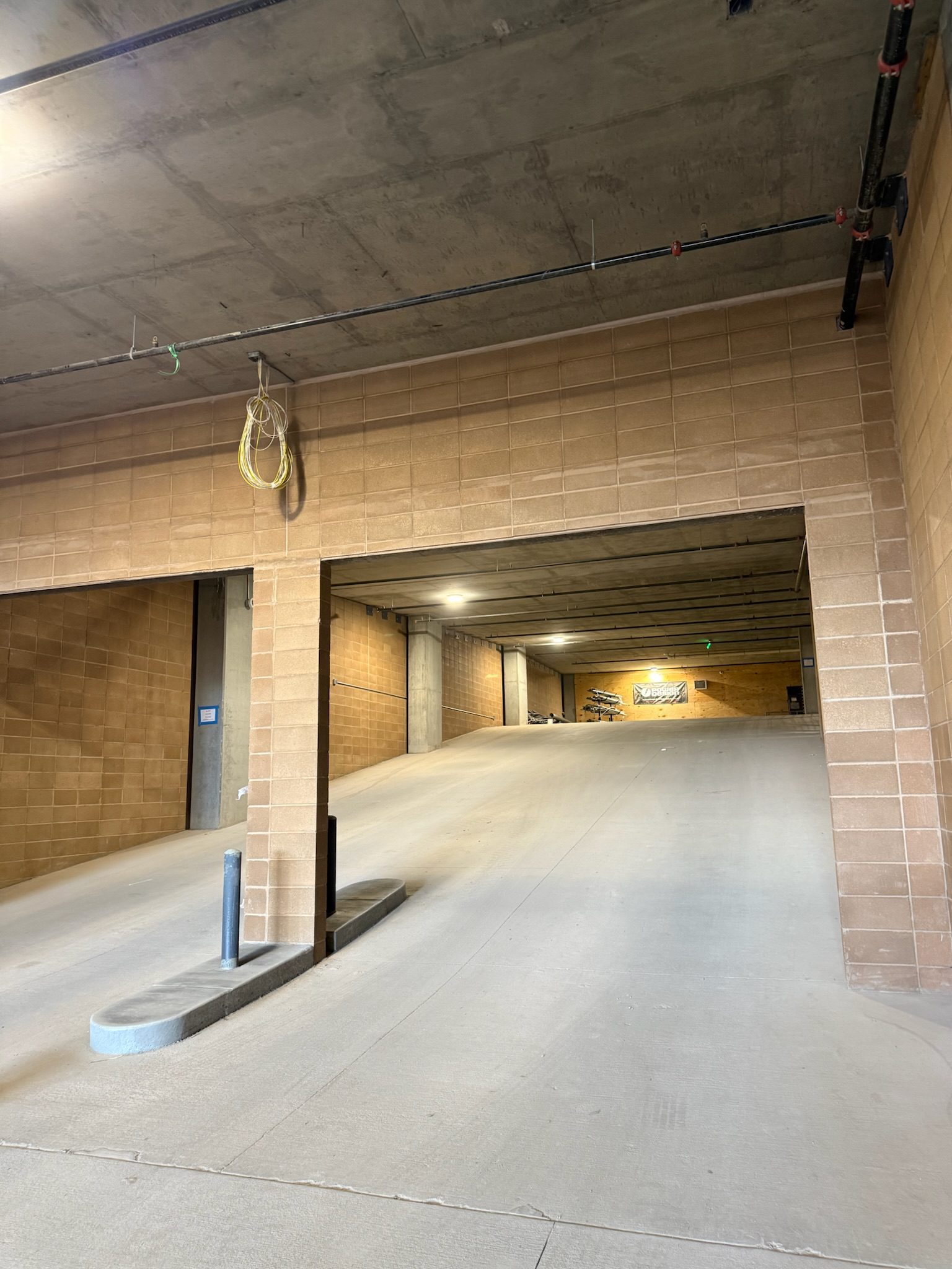 A concrete parking garage ramp with tan brick walls, overhead pipes, and a yellow cord hanging from the ceiling. The ramp leads upward toward brighter light in the distance.