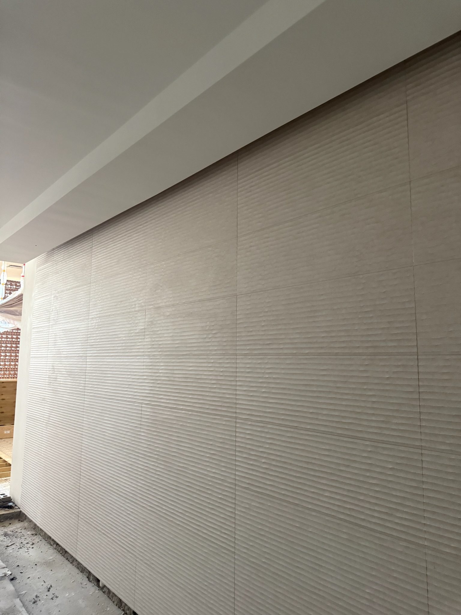 Textured beige tiles are installed on a large interior wall. The floor below the wall is unfinished, with construction materials visible. Light enters from the left side, suggesting ongoing building or renovation work.