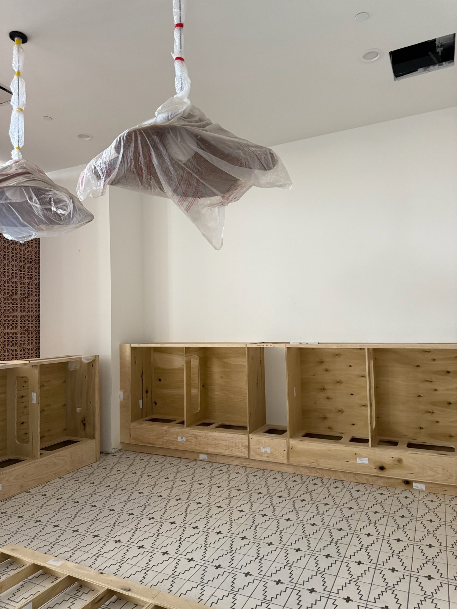 Unfinished room with patterned tile floor, wooden shelving units installed along the wall, and light fixtures hanging from the ceiling covered in plastic. One ceiling tile is missing.